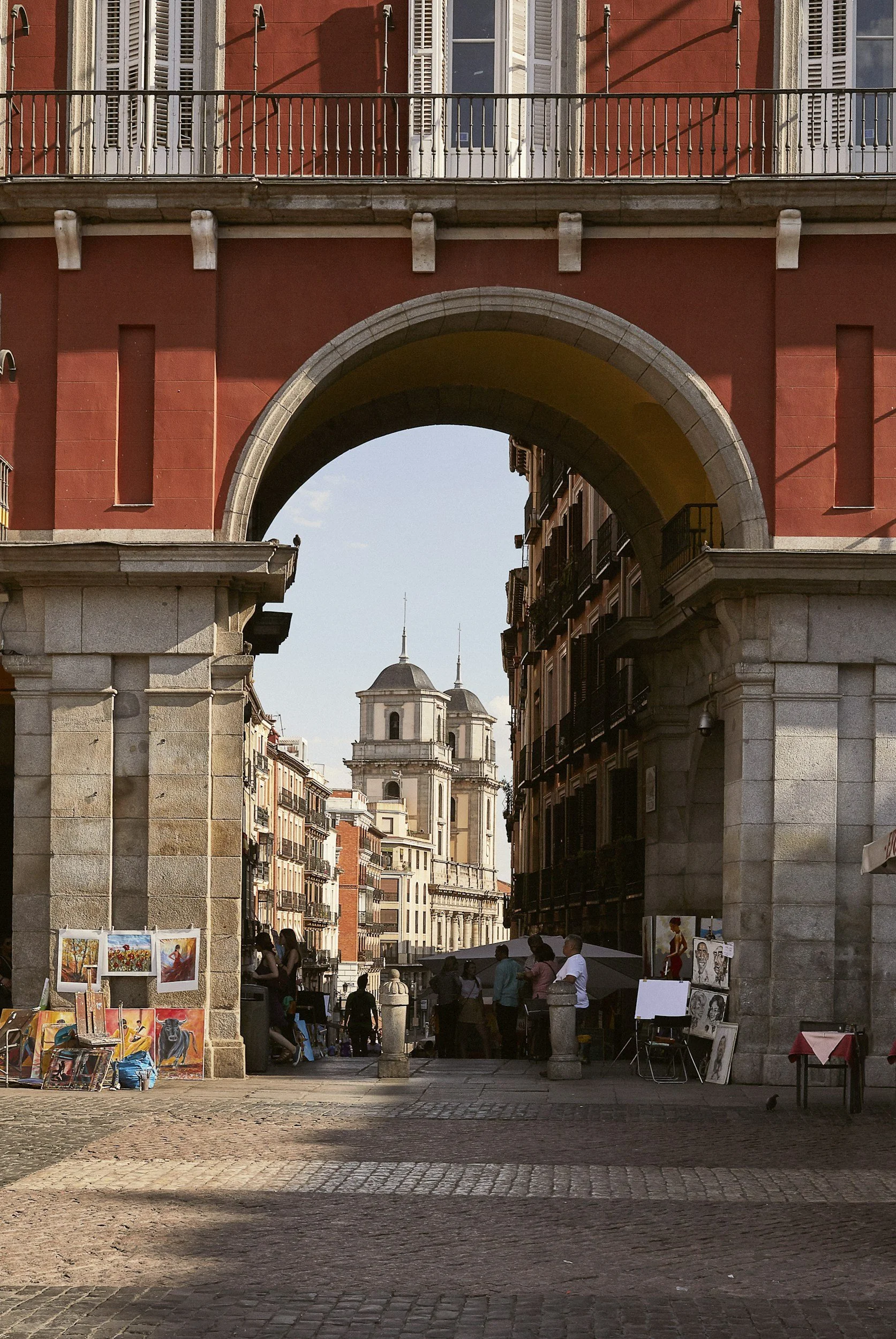 Una escena urbana en un día soleado, con un arco de piedra que enmarca vista hacia un edificio y una iglesia con cúpulas en el fondo, y puesto de arte con pinturas en la calle.
