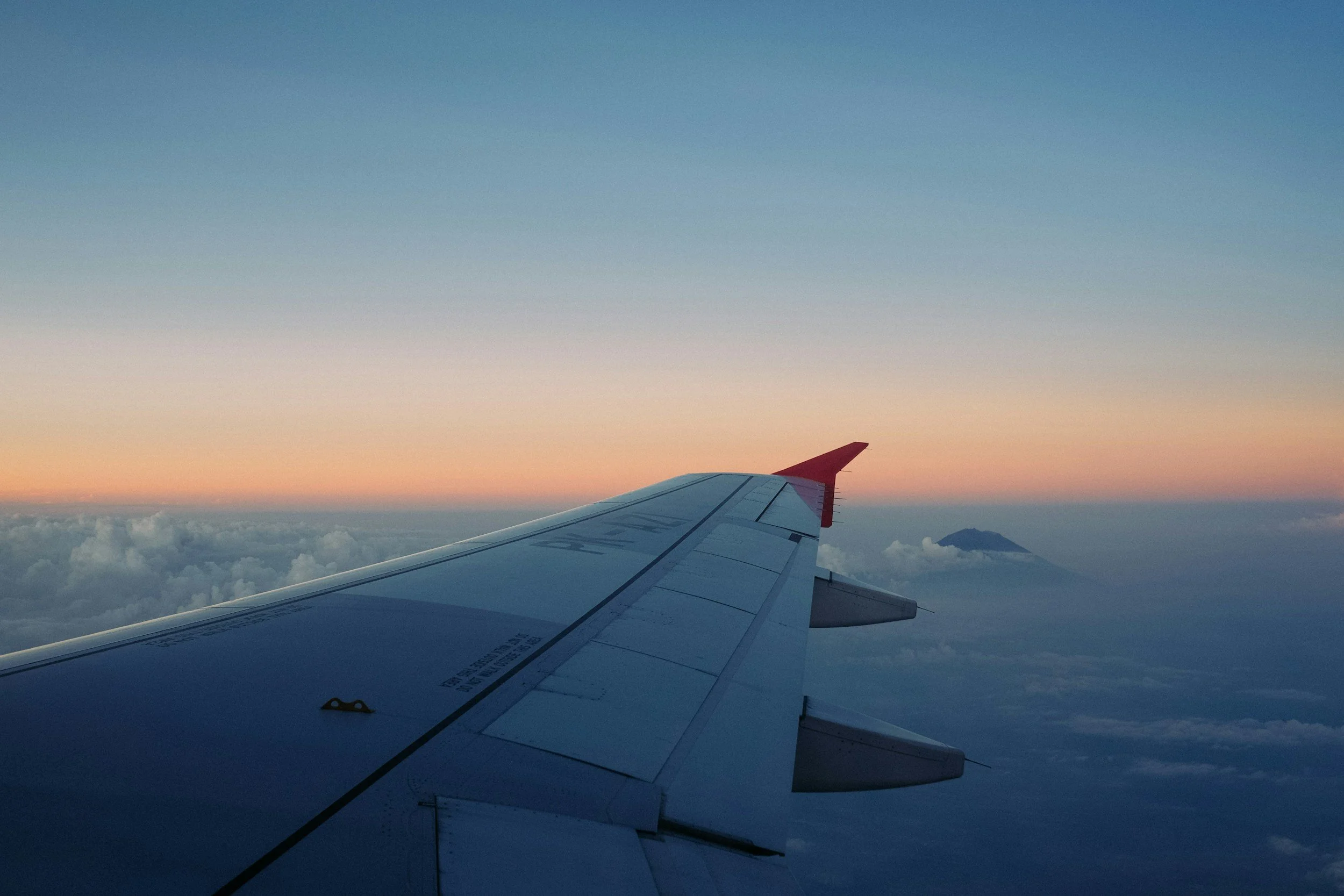 Vista desde la ventana de un avión mostrando una ala, nubes y un volcán en la distancia durante un atardecer o amanecer.