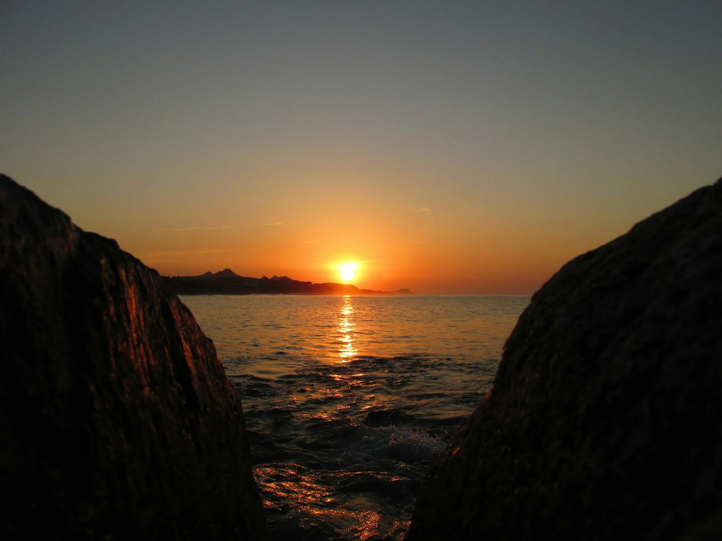Atardecer en la playa con el sol en el horizonte y dos rocas en primer plano.