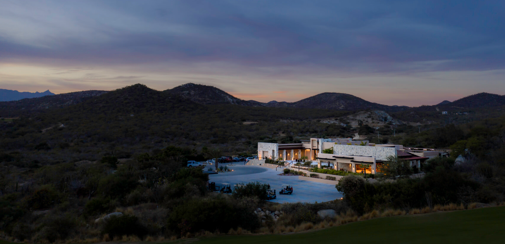 Vista de una casa moderna iluminada en una zona montañosa al atardecer, con un estacionamiento con varios vehículos y un campo de golf.