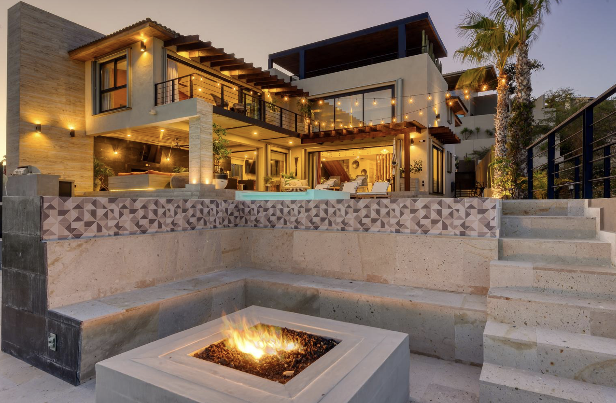 Casa moderna de varios pisos con áreas abiertas, iluminación exterior y mobiliario de patio, con una piscina y fogata en primer plano.