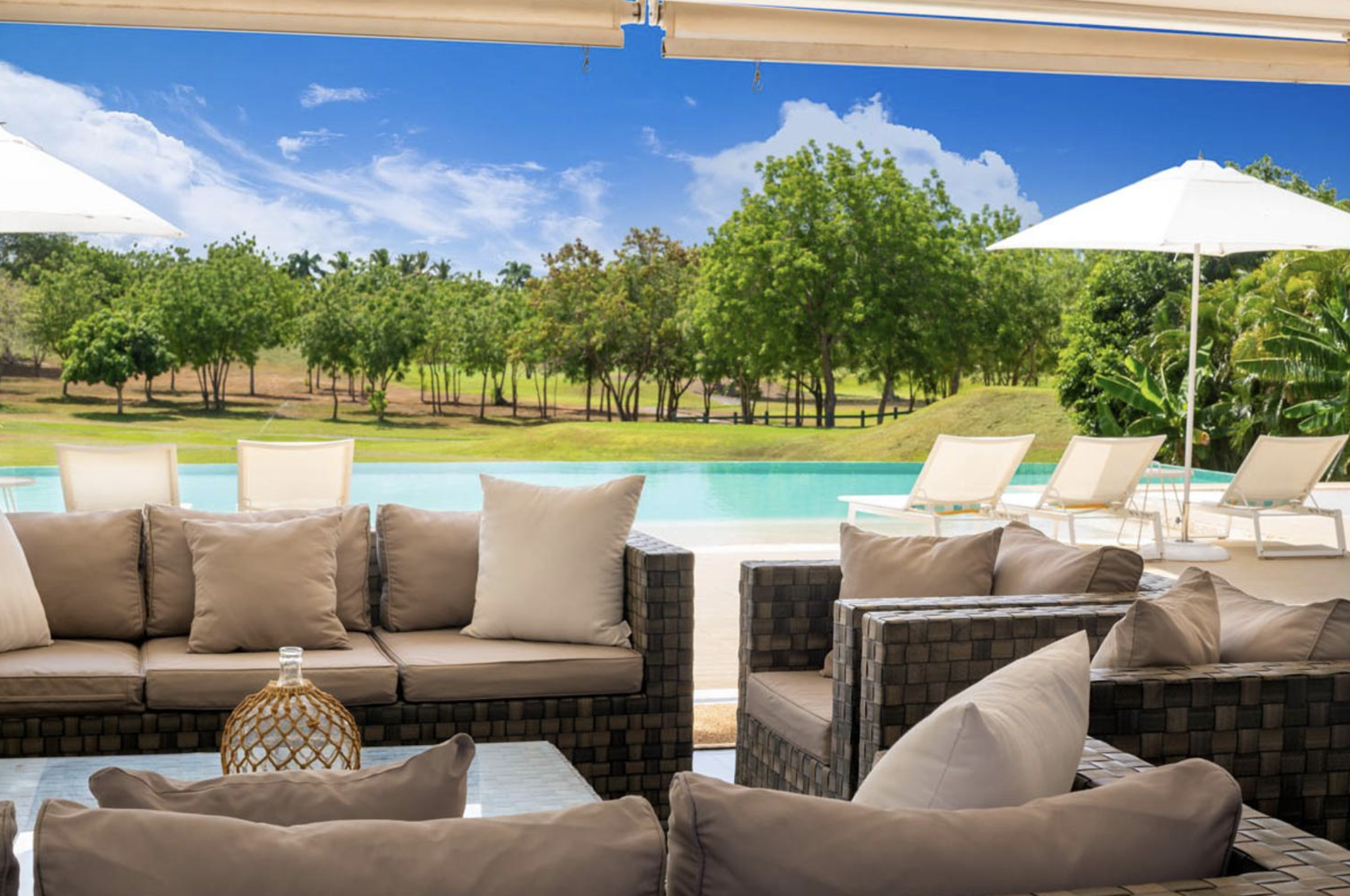 Área de descanso con sillones y cojines, vista a la piscina y áreas verdes con árboles, en un día soleado