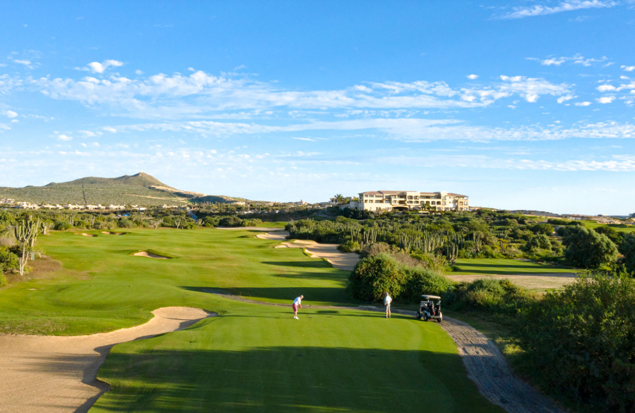 Campo de golf con jugadores y caddy, rodeado de vegetación y edificios al fondo bajo un cielo azul con nubes dispersas.