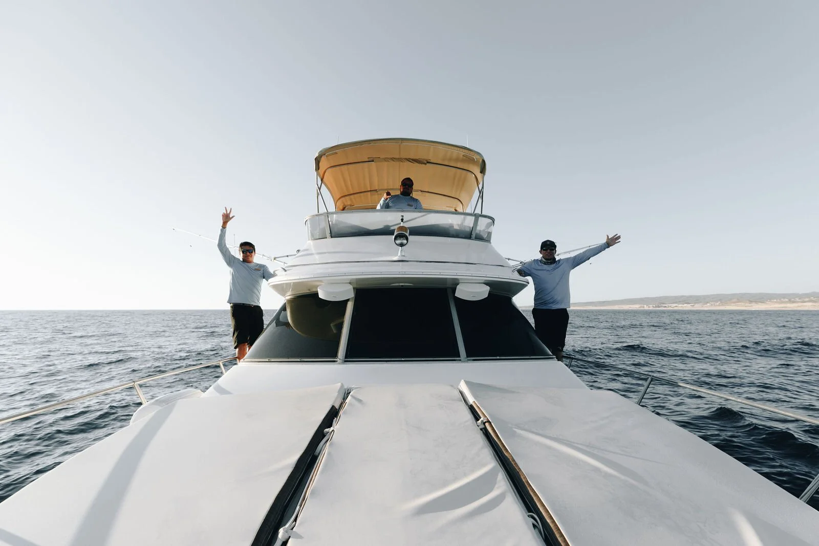Hombres en yate en el mar, uno de ellos en la parte superior con gafas de sol, otros dos en la cubierta con brazos extendidos y saludando.