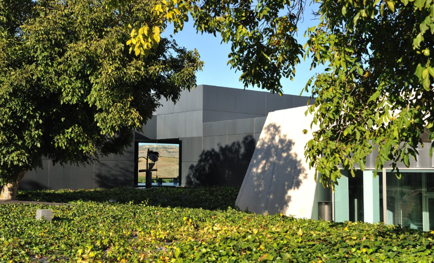 Edificio moderno con paredes negras y blancas, rodeado de árboles y arbustos verdes, bajo un cielo claro y soleado.