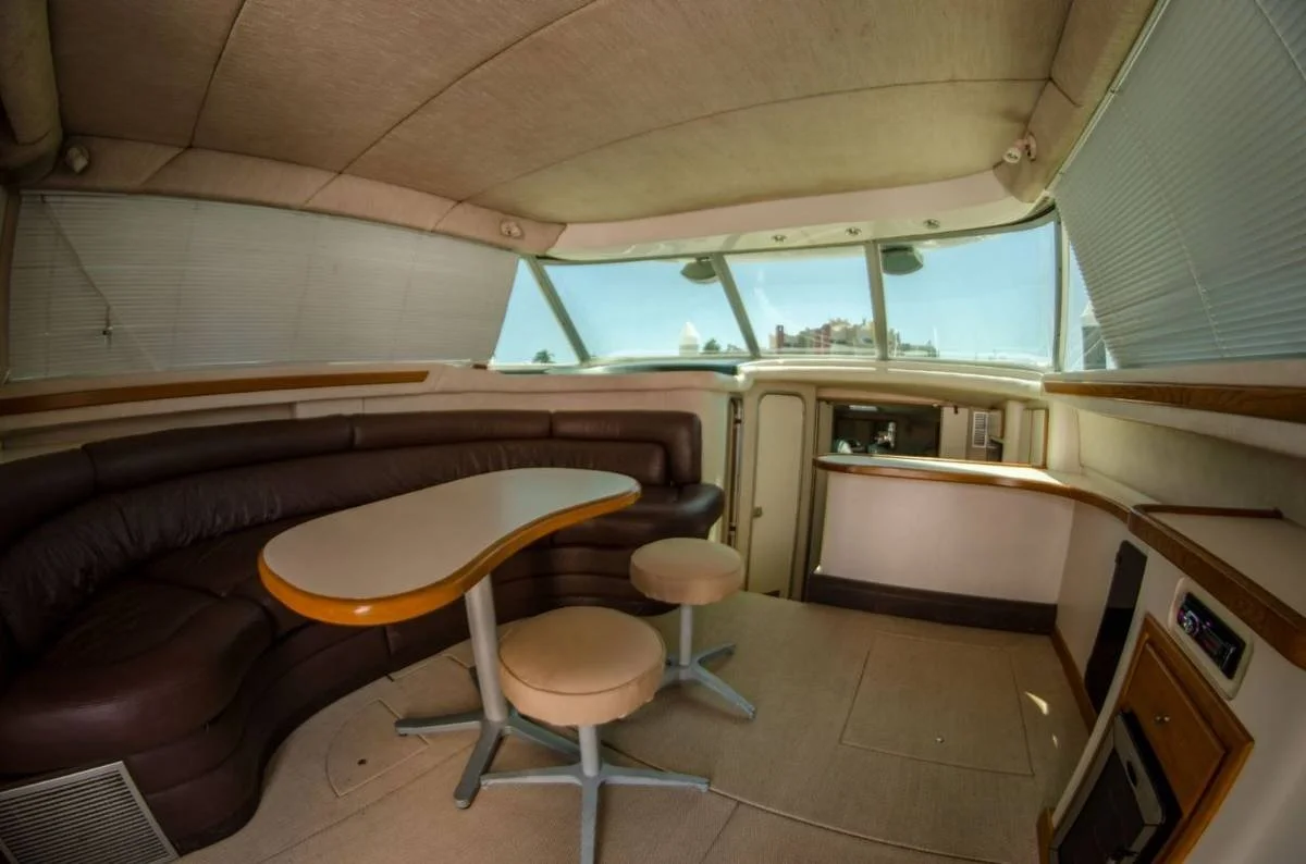 Interior de un yate con sofá en forma de L, mesa ovalada y dos taburetes, ventanas grandes con persianas, y cocina en el fondo.