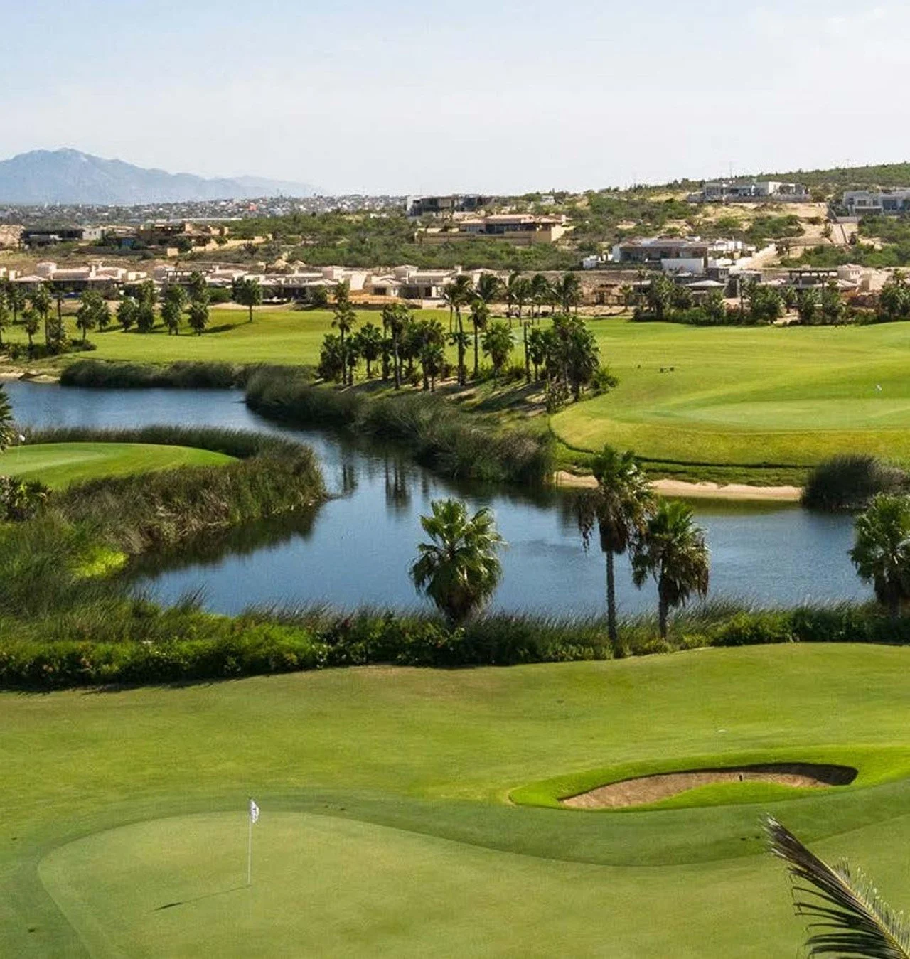 Campo de golf con una laguna y palmeras, vista de casas en las colinas y montañas al fondo.