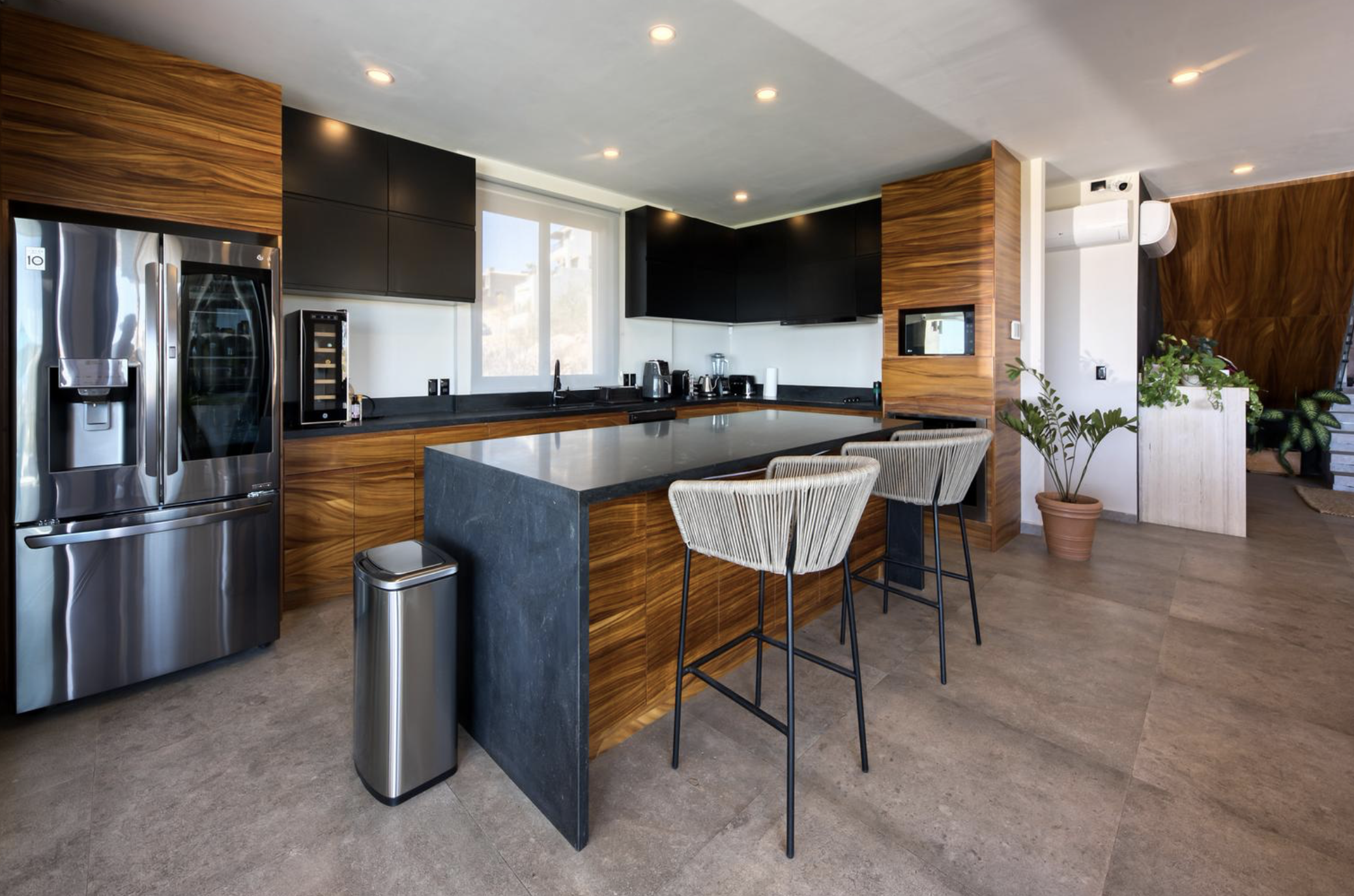 Cocina moderna con nevera de acero inoxidable, encimera negra, gabinetes de madera y paredes blancas, con bar con dos sillas de respaldo de cuerda y plantas decorativas.