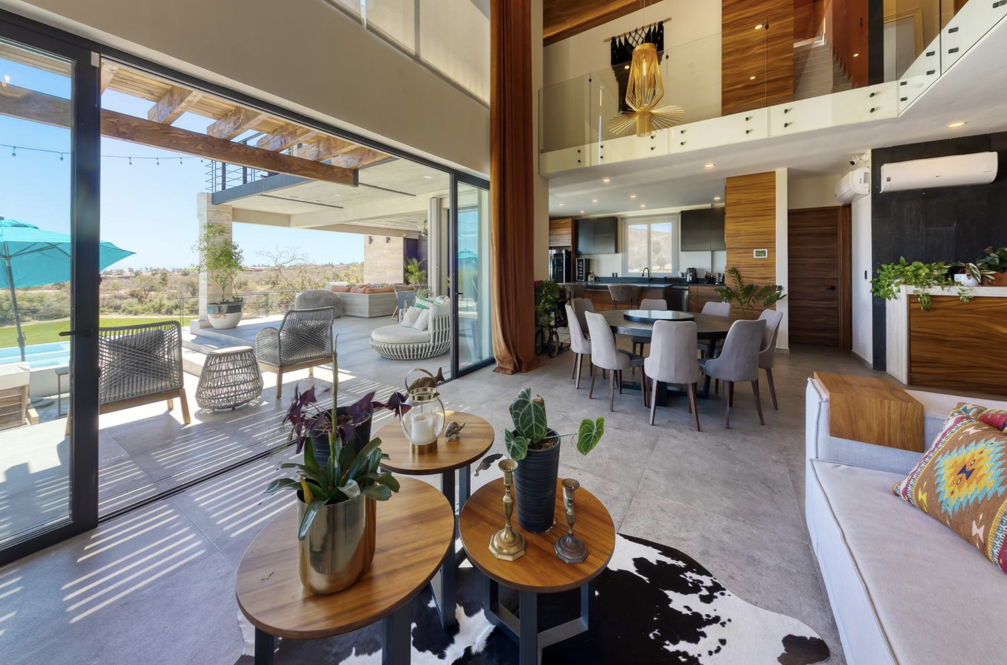 Interior de una casa moderna con sala de estar y comedor, con vista a una terraza exterior con mobiliario y áreas verdes cercanas.