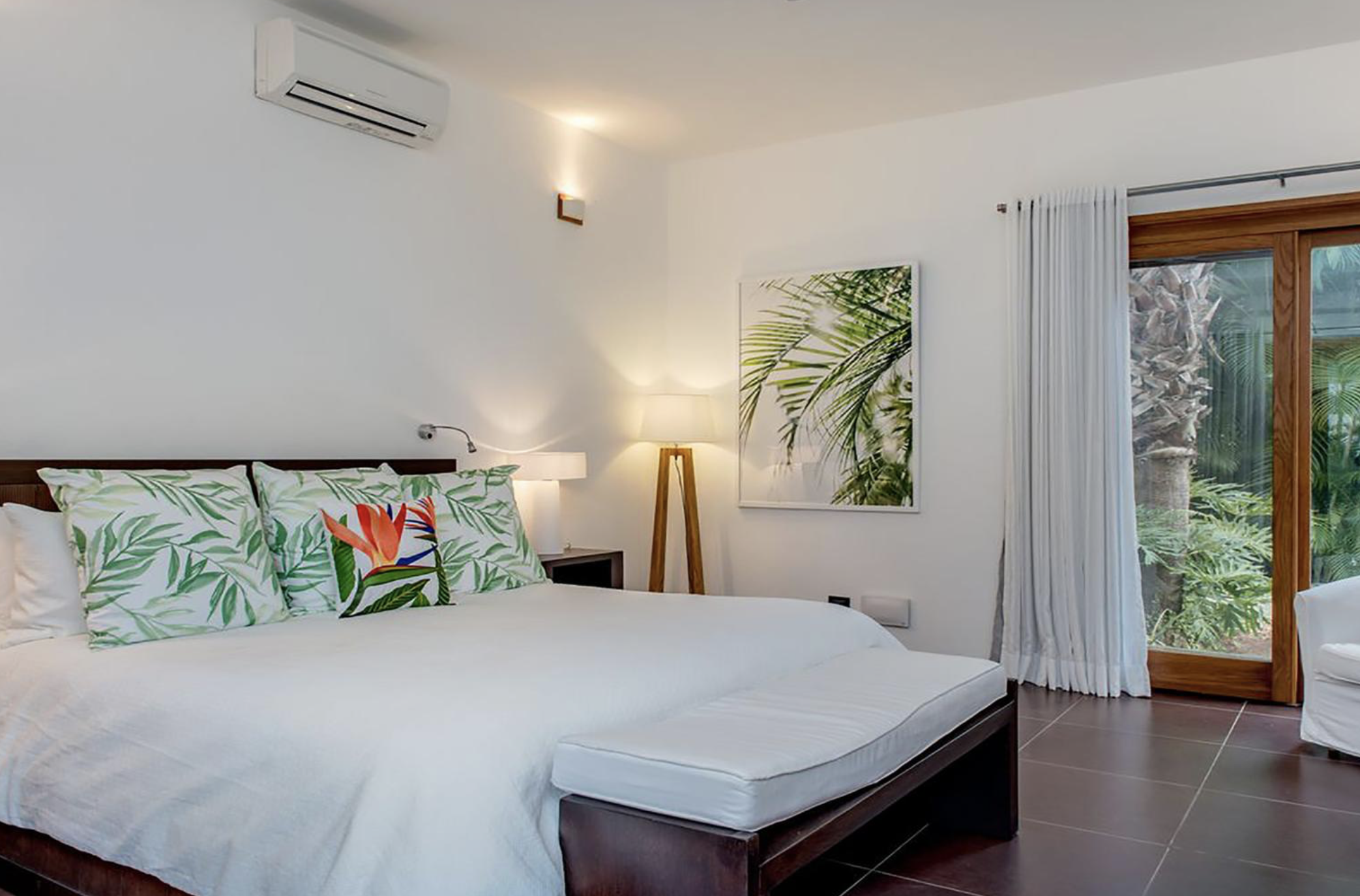 Habitación moderna con cama grande, almohadas con funda de diseño de hojas verdes y flor tropical, lámpara de pie, cuadro con motivo de hojas, vista a jardín desde ventana de madera y cortinas blancas.