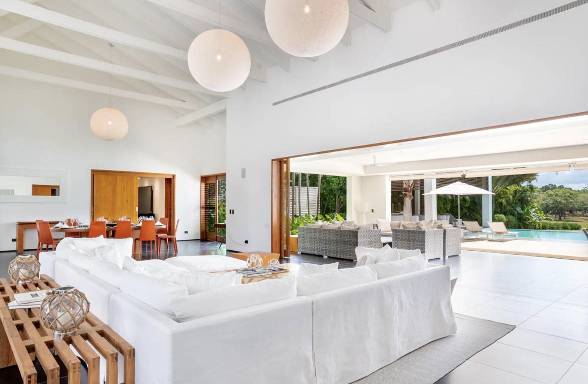 Sala de estar moderna con mueble blanco, comedor con sillas naranjas, espacio abierto con vista a la piscina y jardín exterior.