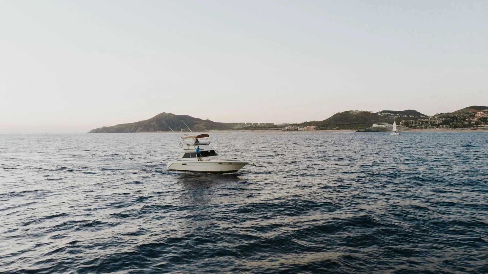 Barco de recreo en el mar con colina y paisaje urbano en el fondo, cielo despejado.