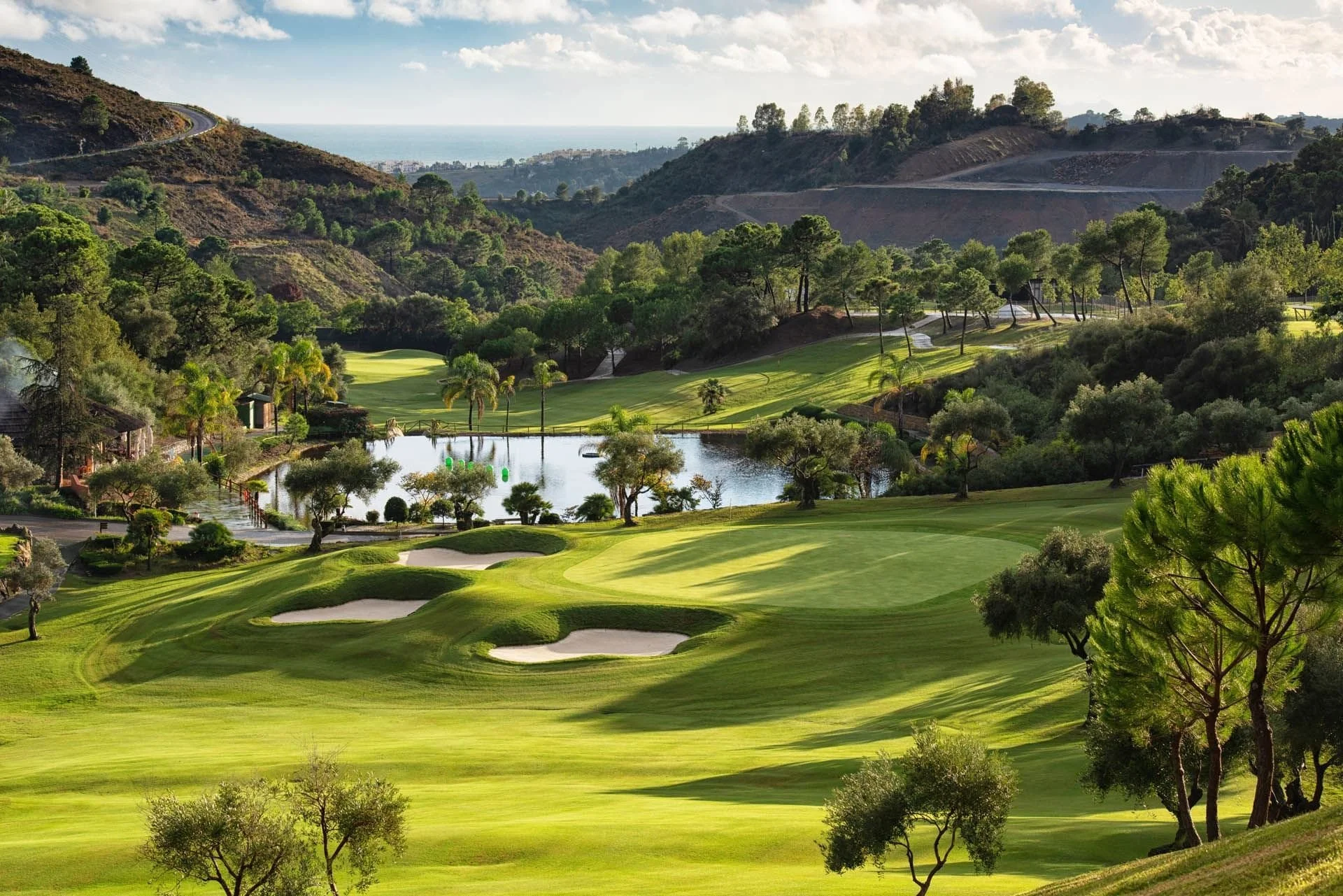 Campo de golf con áreas verdes, árboles y un lago, rodeado de colinas y montañas, con vista al mar en el fondo.
