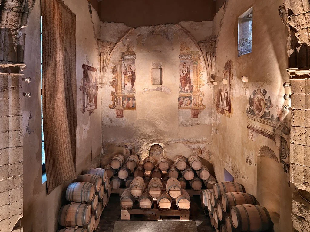 Sala de bodegas con barriles de vino apilados y paredes decoradas con frescos religiosos antiguos.