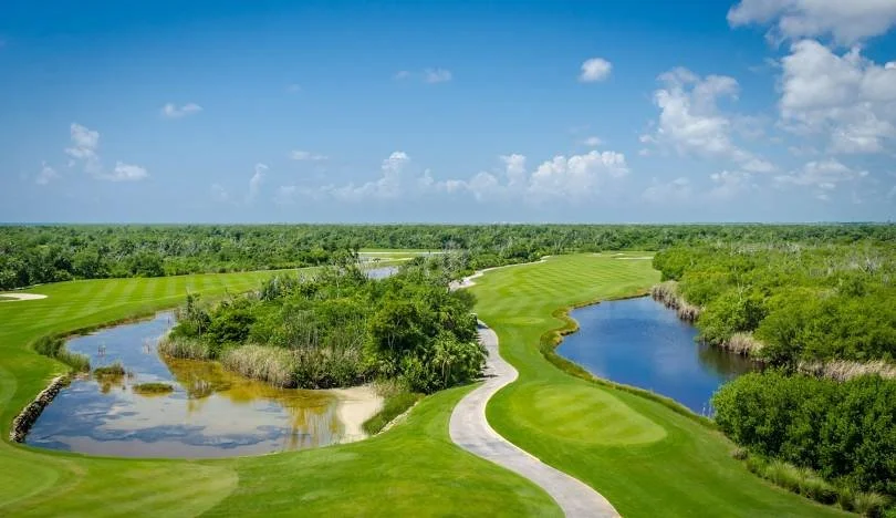 Campo de golf con caminos, lagos y vegetación, bajo un cielo parcialmente nublado.