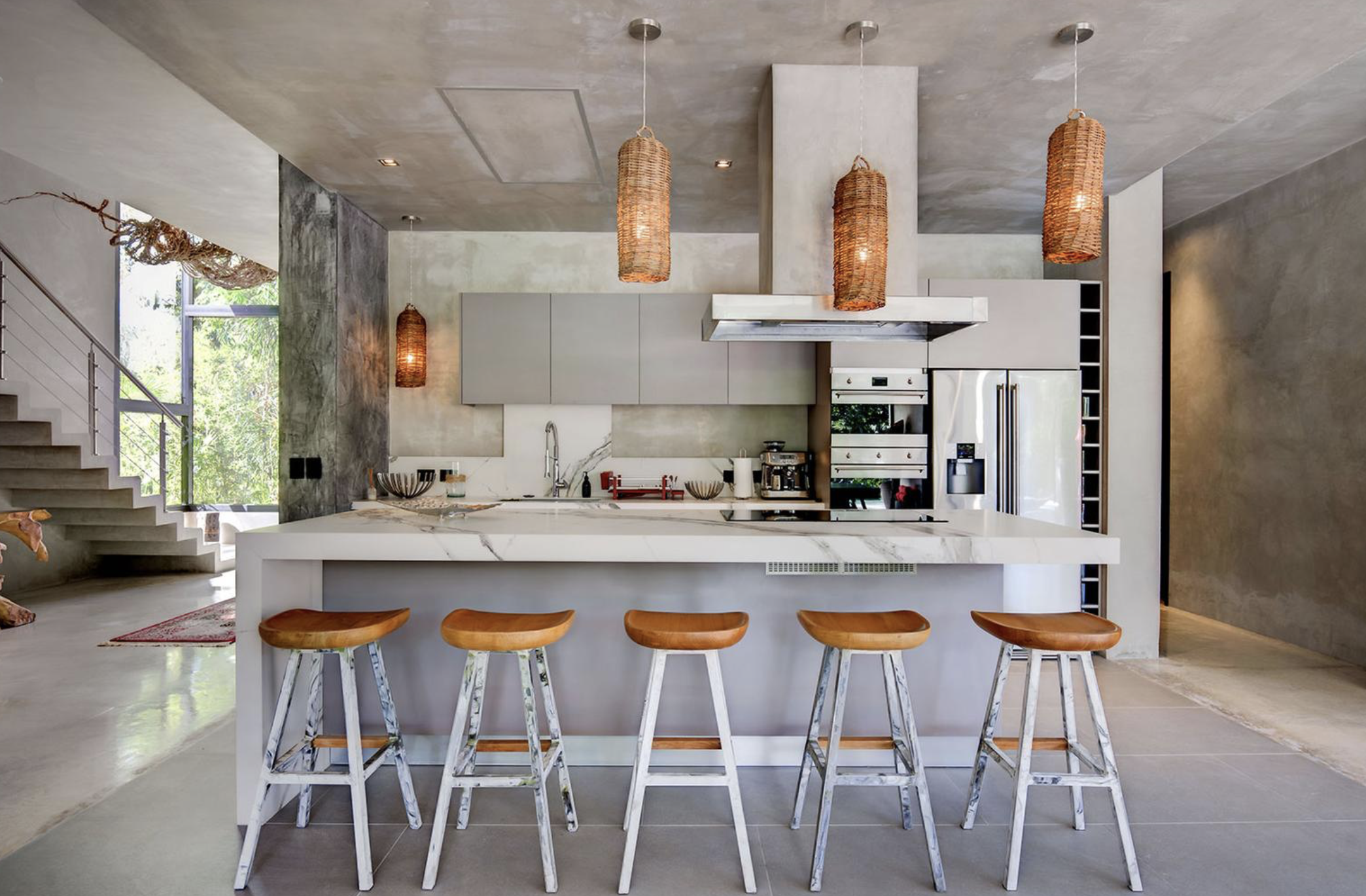 Cocina moderna con isla de mármol, cinco sillas con asientos de madera y bases de metal, pendant lights de mimbre, electrodomésticos de acero inoxidable y ventanas grandes con vista a árboles.