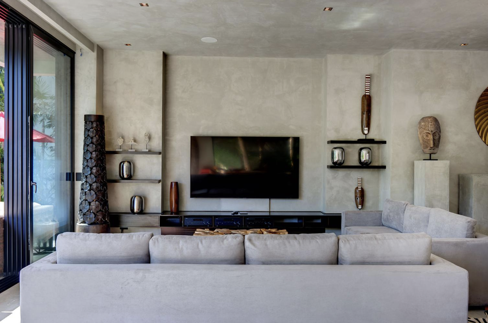 Sala de estar moderna con un televisor grande en la pared, sillones claros, estanterías con decoraciones y esculturas de madera, y paredes de color beige.