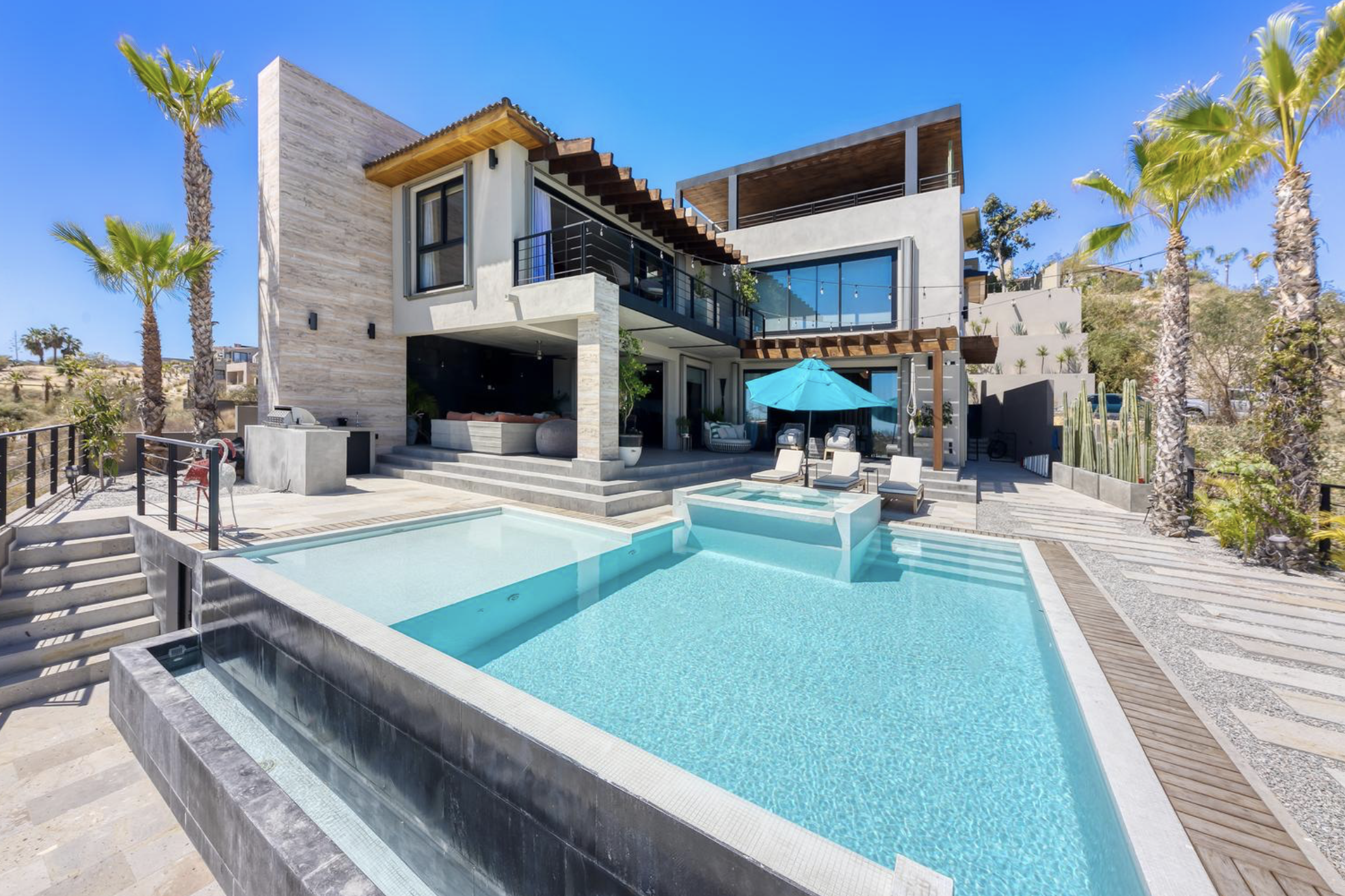 Casa moderna con piscina y jardín en un día soleado, palmeras y terraza al aire libre.