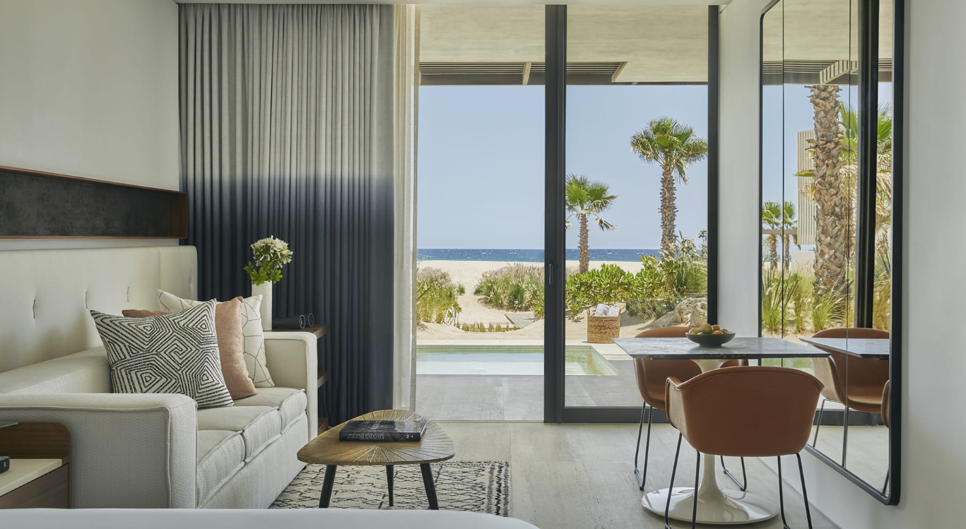 Sala con vista al mar y a la playa, con sillón, mesa de café, televisor, mesa redonda con sillas, y vista a palmeras y arena desde ventanas grandes.
