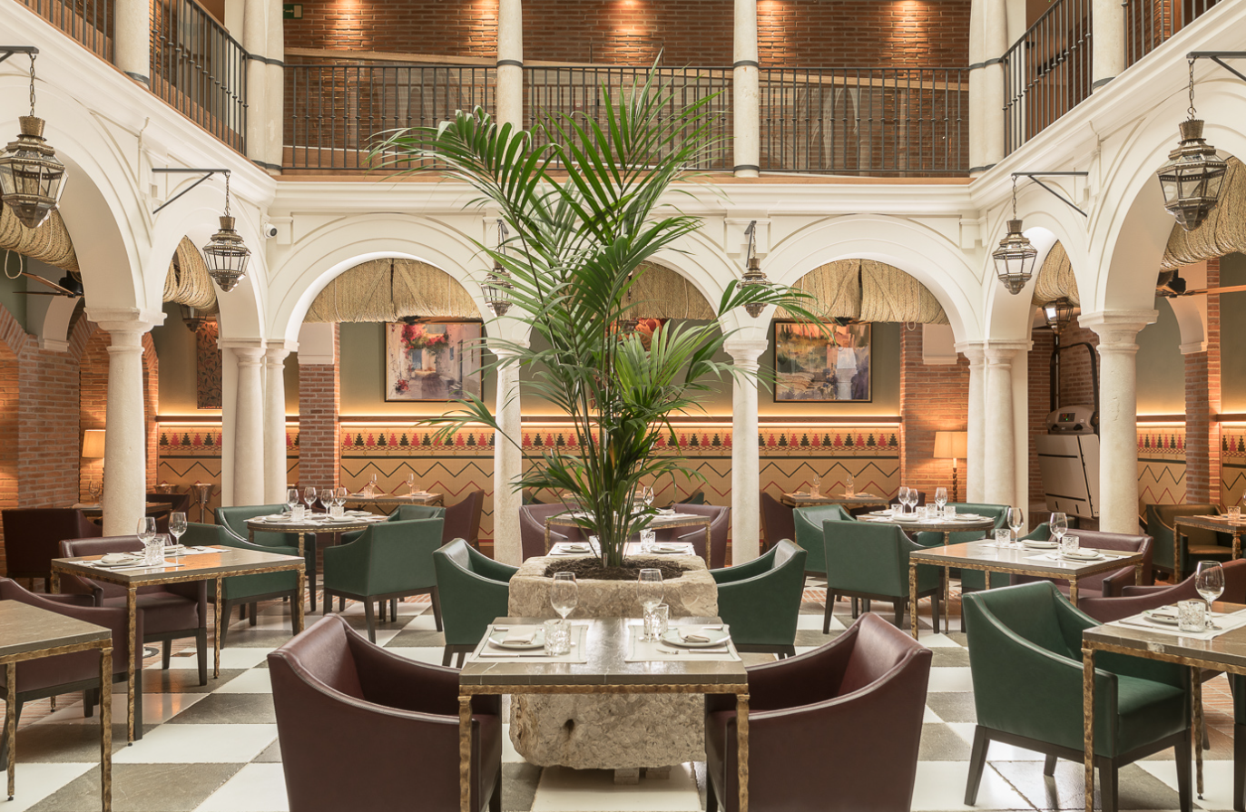 Interior elegante de restaurante con mesas decoradas, sillas de color marrón y verde, una planta grande en el centro y paredes de ladrillo y arcos blancos.