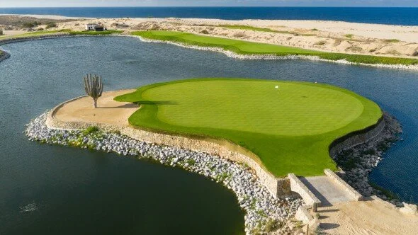 Vista aérea de un campo de golf en medio de un lago artificial, cerca de la playa.