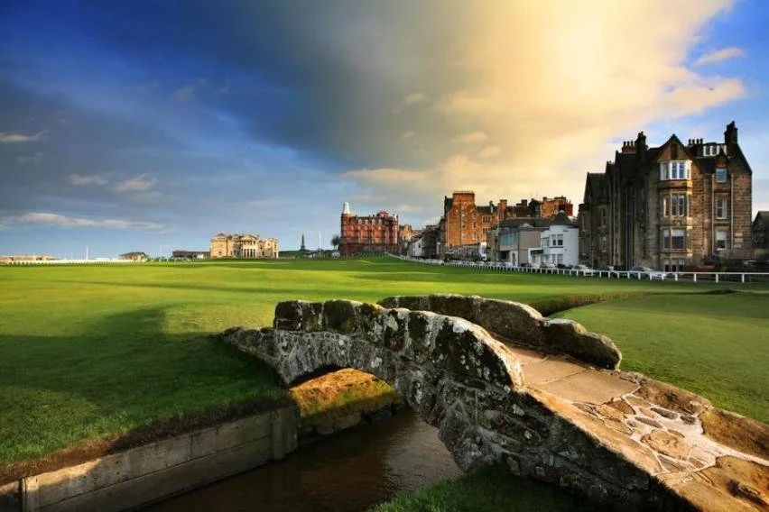 Campo de golf con puente de piedra y edificios históricos al fondo, bajo un cielo con nubes y sol.