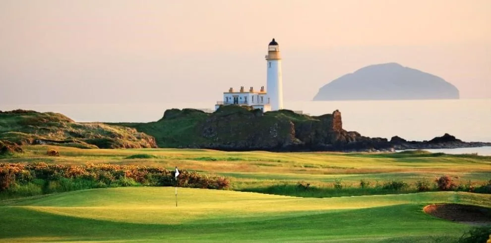 Un campo de golf con un faro en la isla cercana y el mar al fondo.