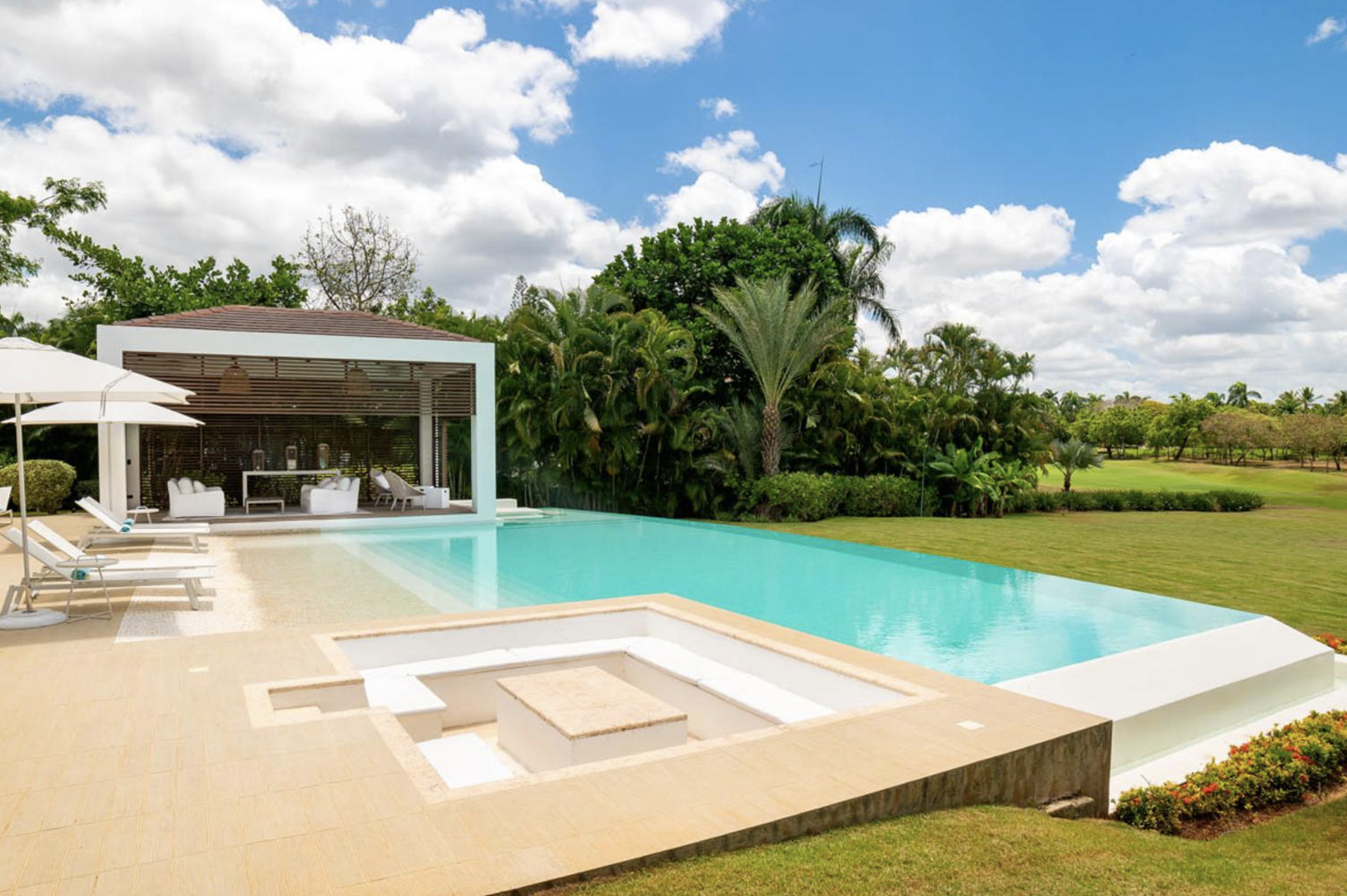 Piscina moderna en un espacio exterior con césped, sombrillas y tumbonas, rodeada de árboles verdes y un cielo con nubes.