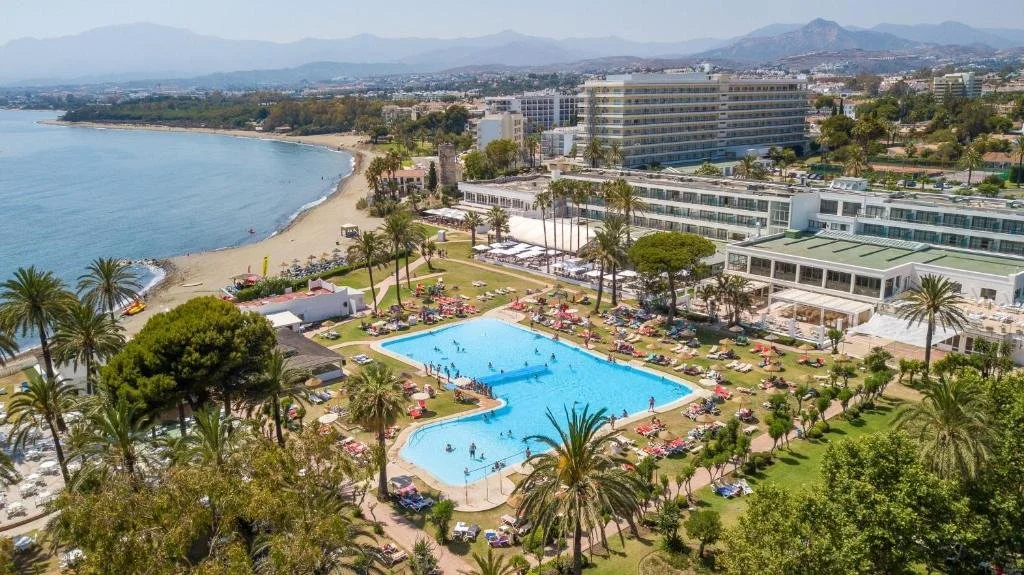 Vista aérea de un complejo turístico con piscina, palmeras, una playa y edificios en un entorno costero rodeado de montañas.