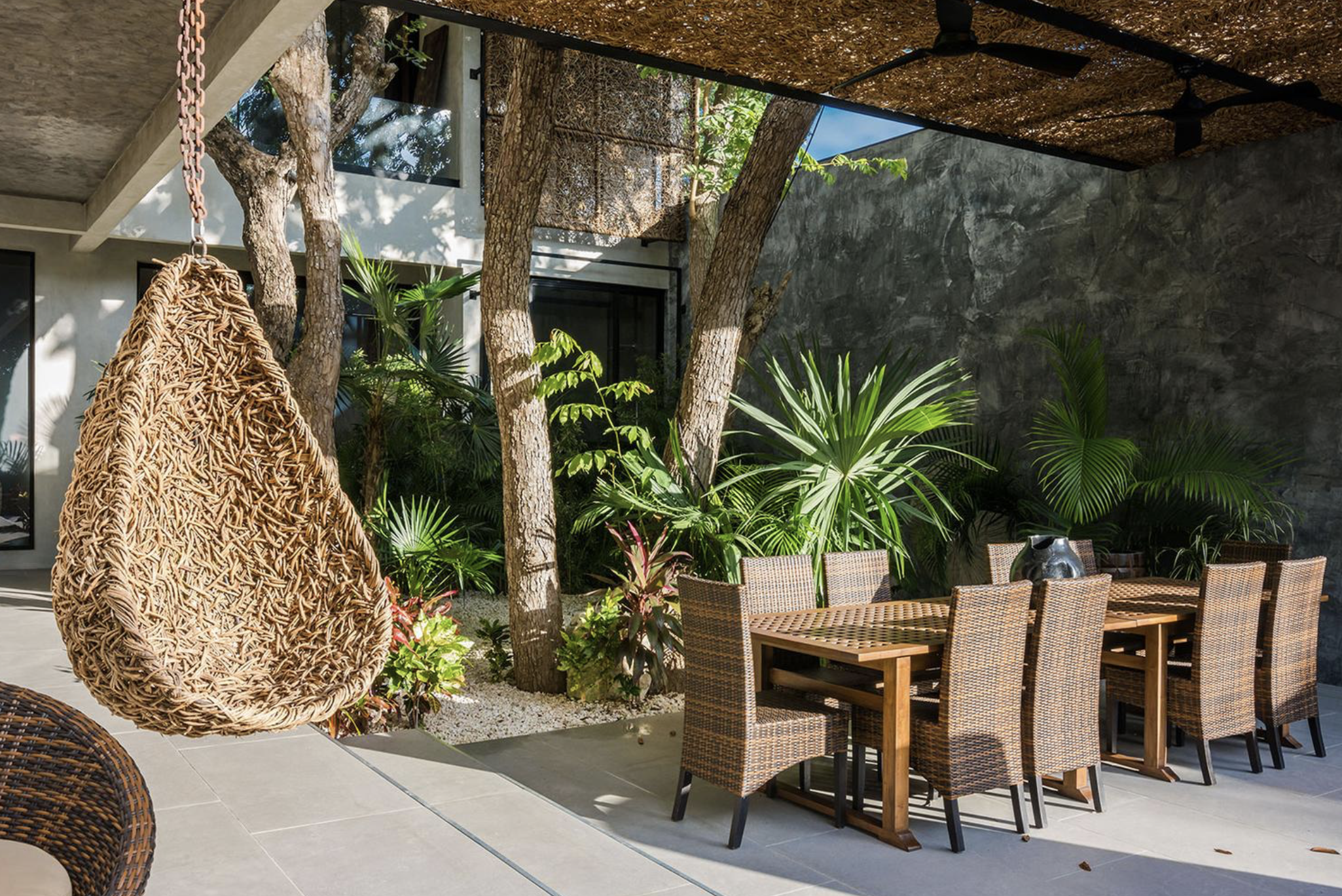 Área de comedor al aire libre con mesas y sillas de mimbre, rodeada de plantas tropicales y árboles, con pared de piedra y techos con ventiladores de techo.