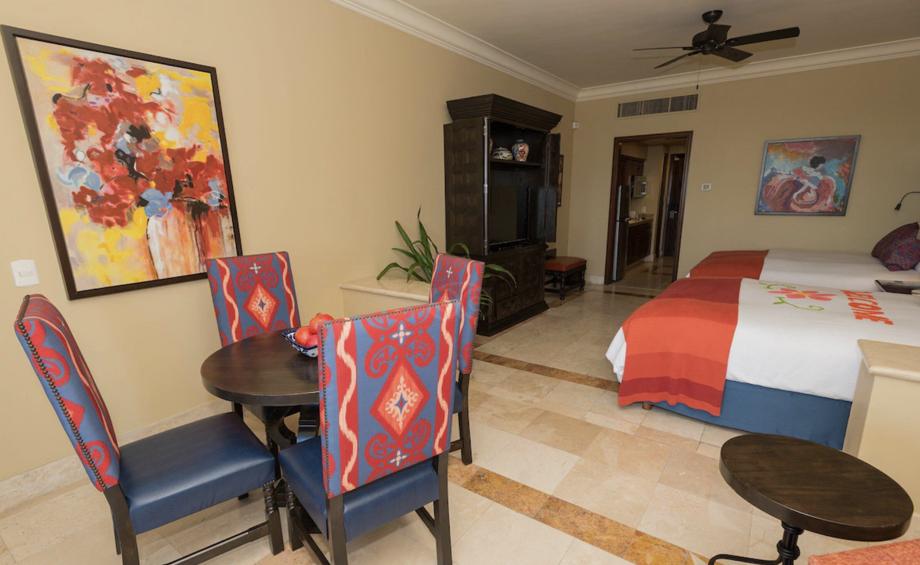 Habitación de hotel con dos camas, una mesa redonda con cuatro sillas tapizadas, armario con televisor, obras de arte en las paredes, y piso de baldosas beige.