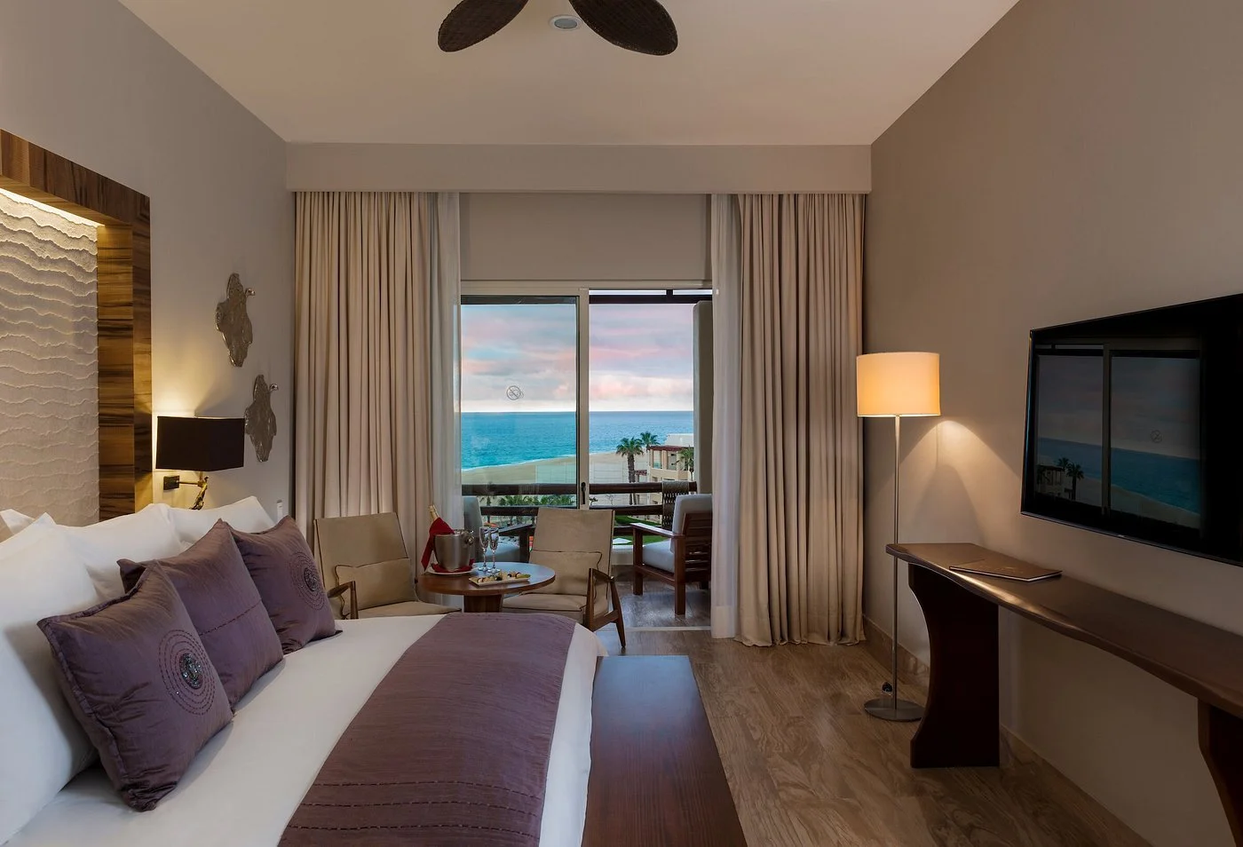 Habitación de hotel con vista al mar, cama con almohadas moradas y cortinas blancas.