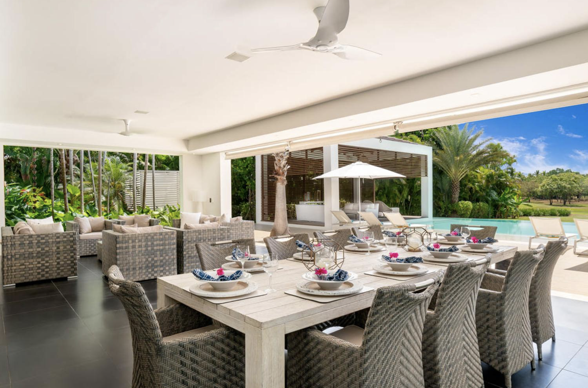Comedor al aire libre con mesa y sillas, vista hacia la piscina, terraza y vegetación tropical.