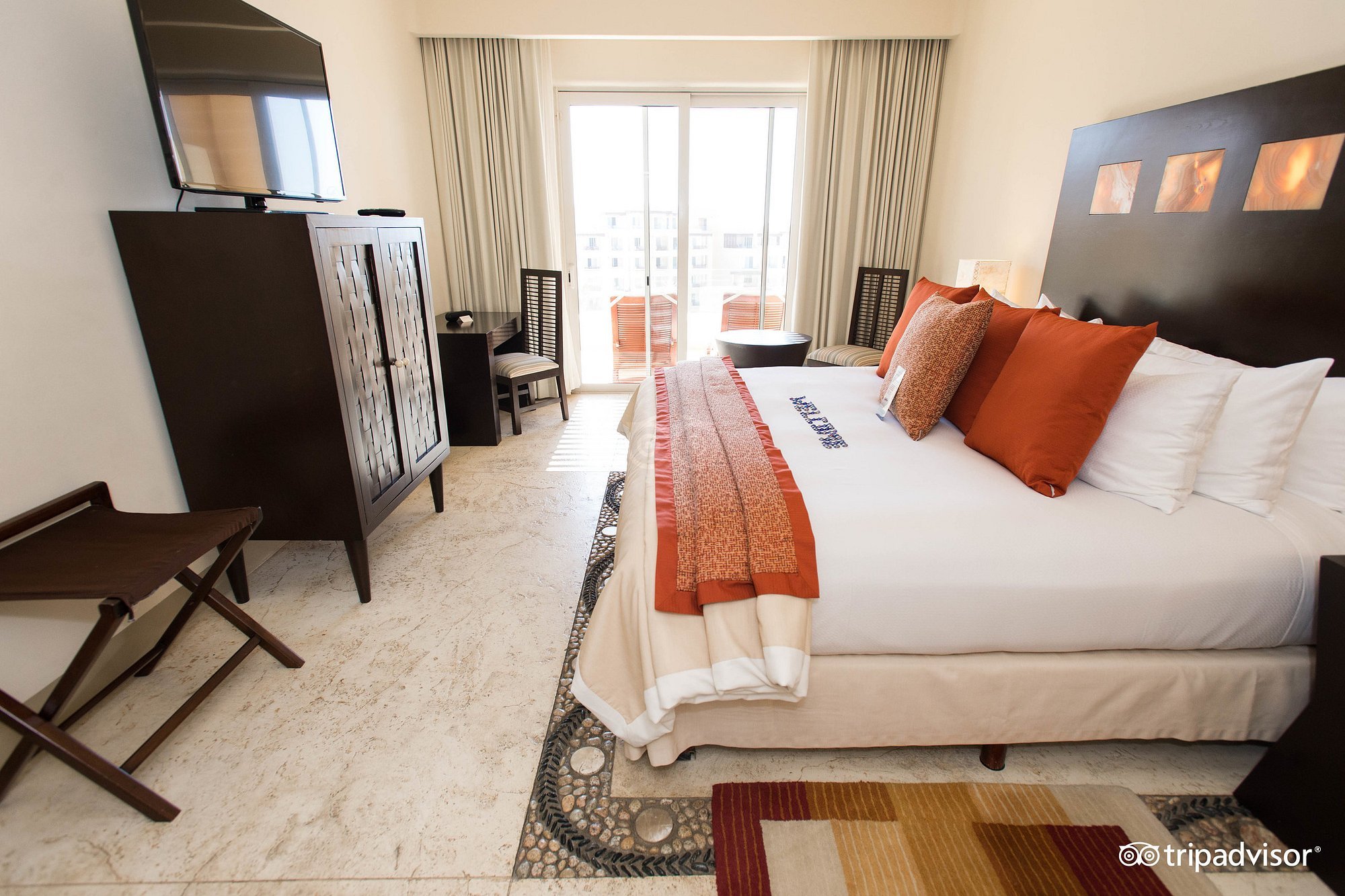 Habitación de hotel con cama grande con almohadas de color naranja y beige, televisión en mueble oscuro, ventana con cortinas, silla de madera, y suelo de piedra con alfombra rectangular.