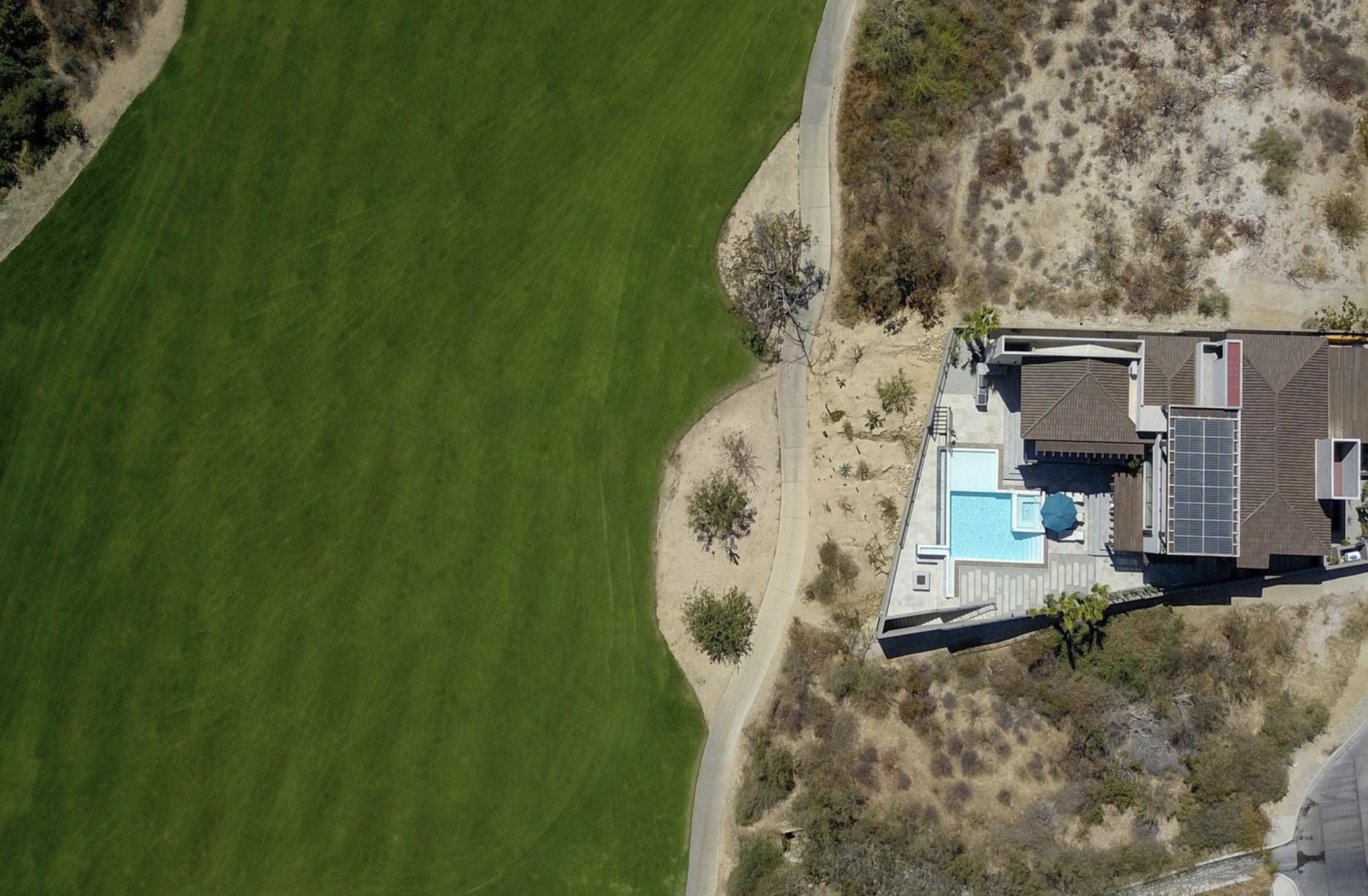 Vista aérea de una casa con piscina y paneles solares en un vecindario, rodeada de áreas verdes y terreno árido.