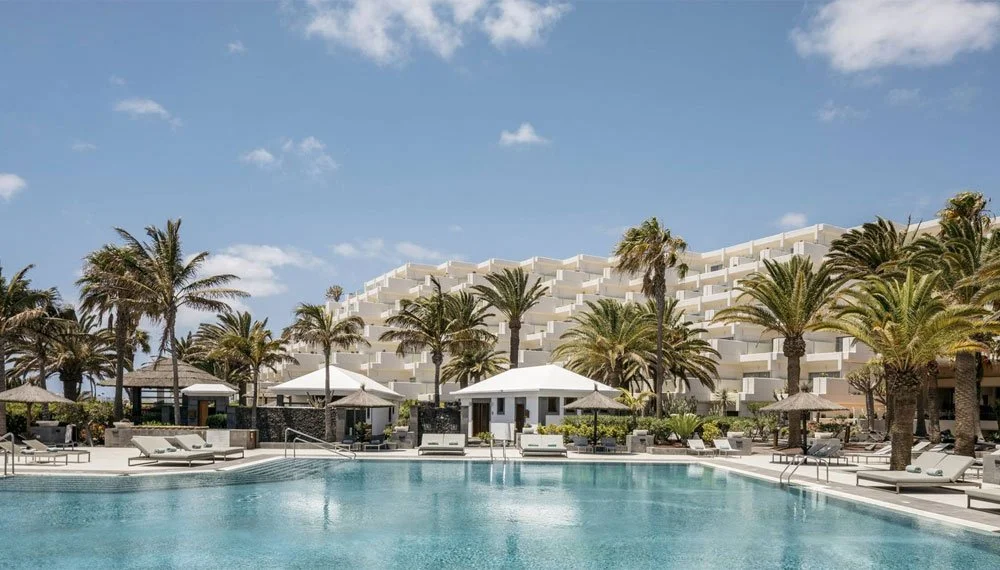 Vista de una alberca en un resort con palmeras y construcciones blancas al fondo, cielo azul con algunas nubes