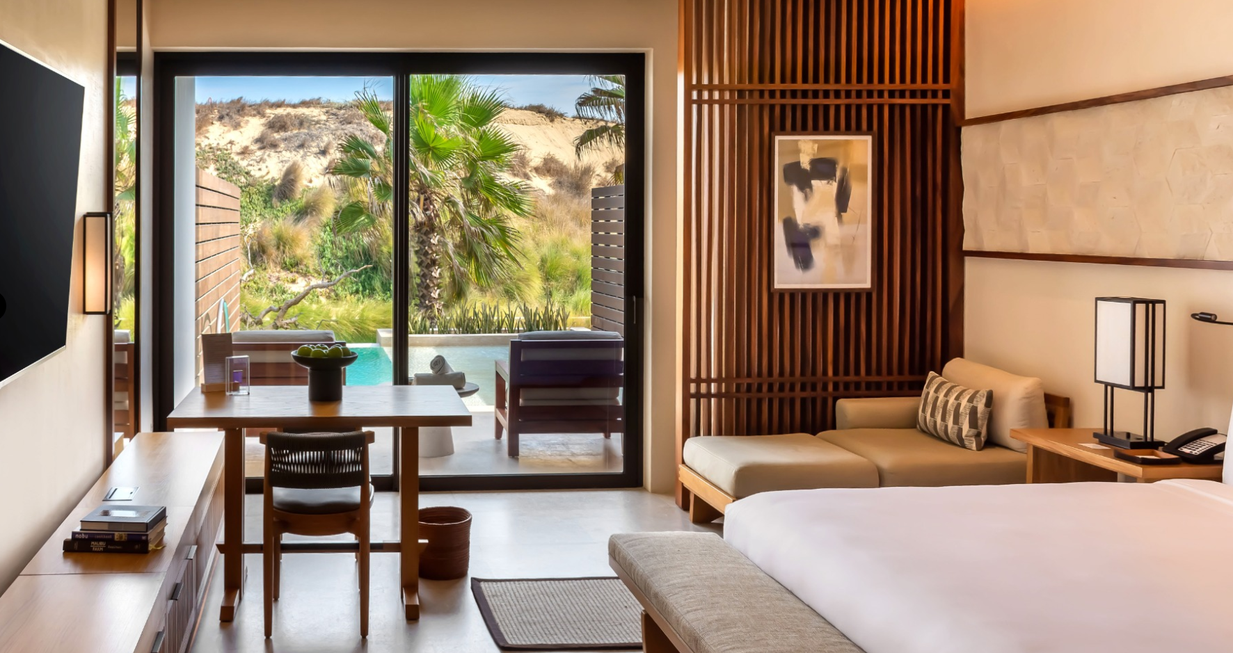 Habitación con vista a un jardín con palmeras y una piscina, muebles de madera, cama, sillón y mesa con lámpara decorativa.