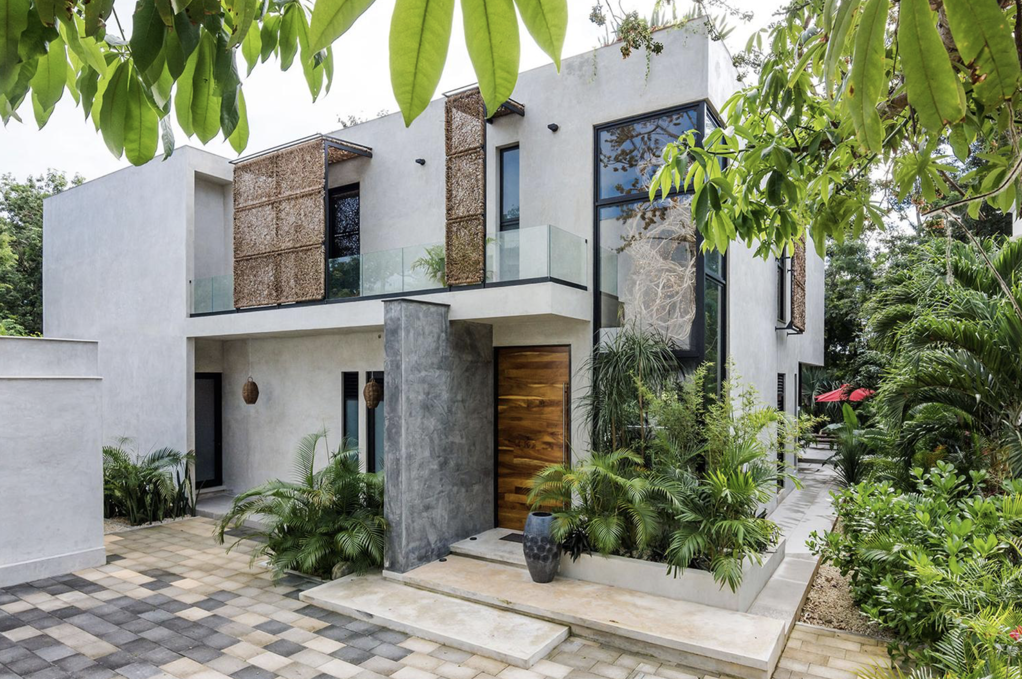 Casa moderna con paredes blancas y grandes ventanales, rodeada de vegetación y plantas tropicales. Entrada principal con puertas de madera y macetas decorativas.