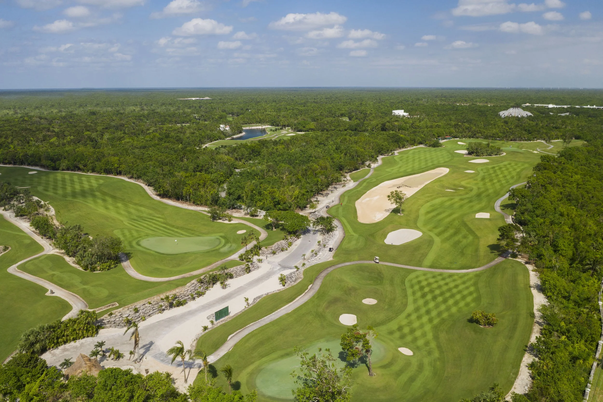 Campo de golf con césped bien cuidado, algunos bunkers y árboles dispersos, rodeado de vegetación y un cielo parcialmente nublado.
