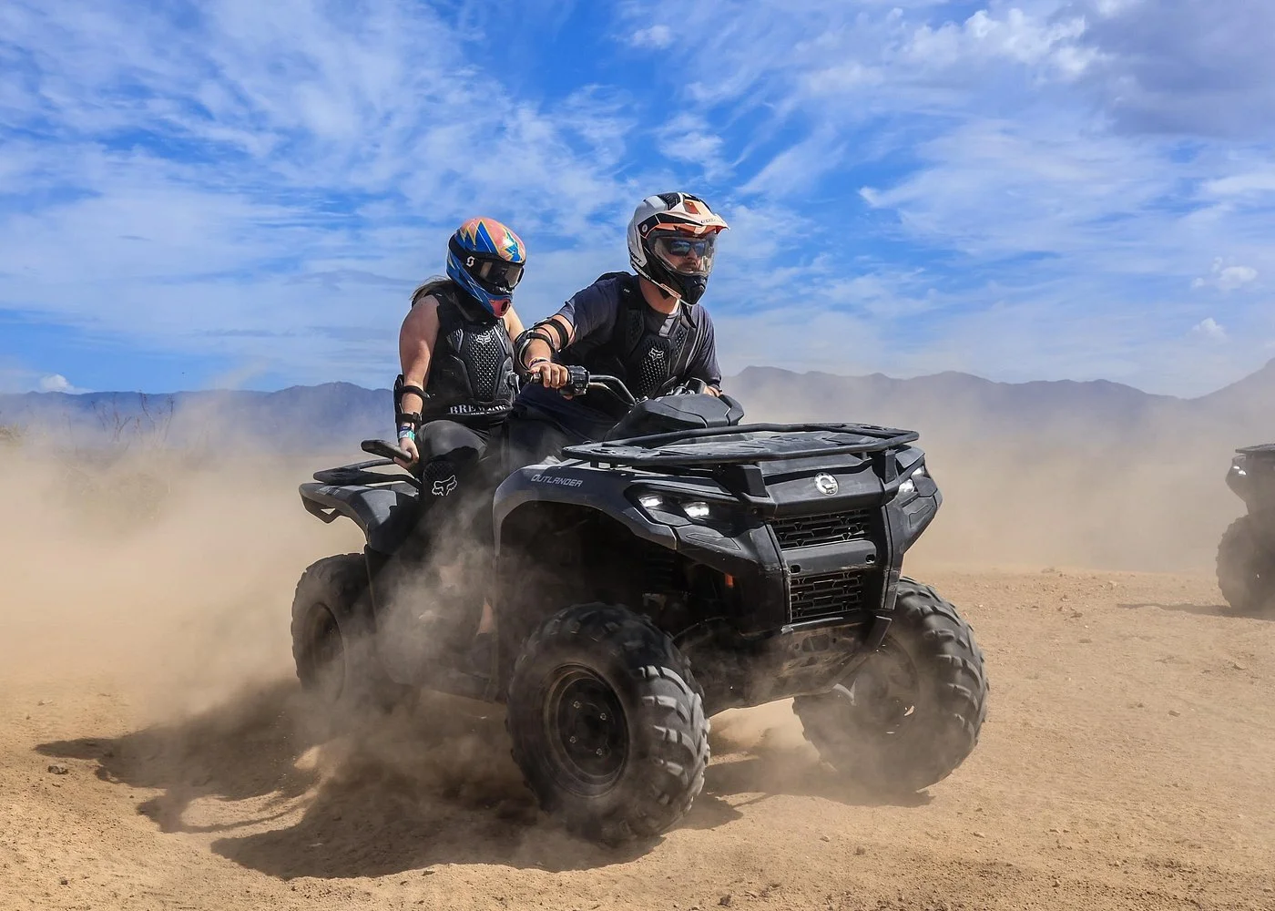 Dos personas en cascos y ropa de protección conduciendo un vehículo todo terreno en un paisaje árido con polvo y montañas al fondo.