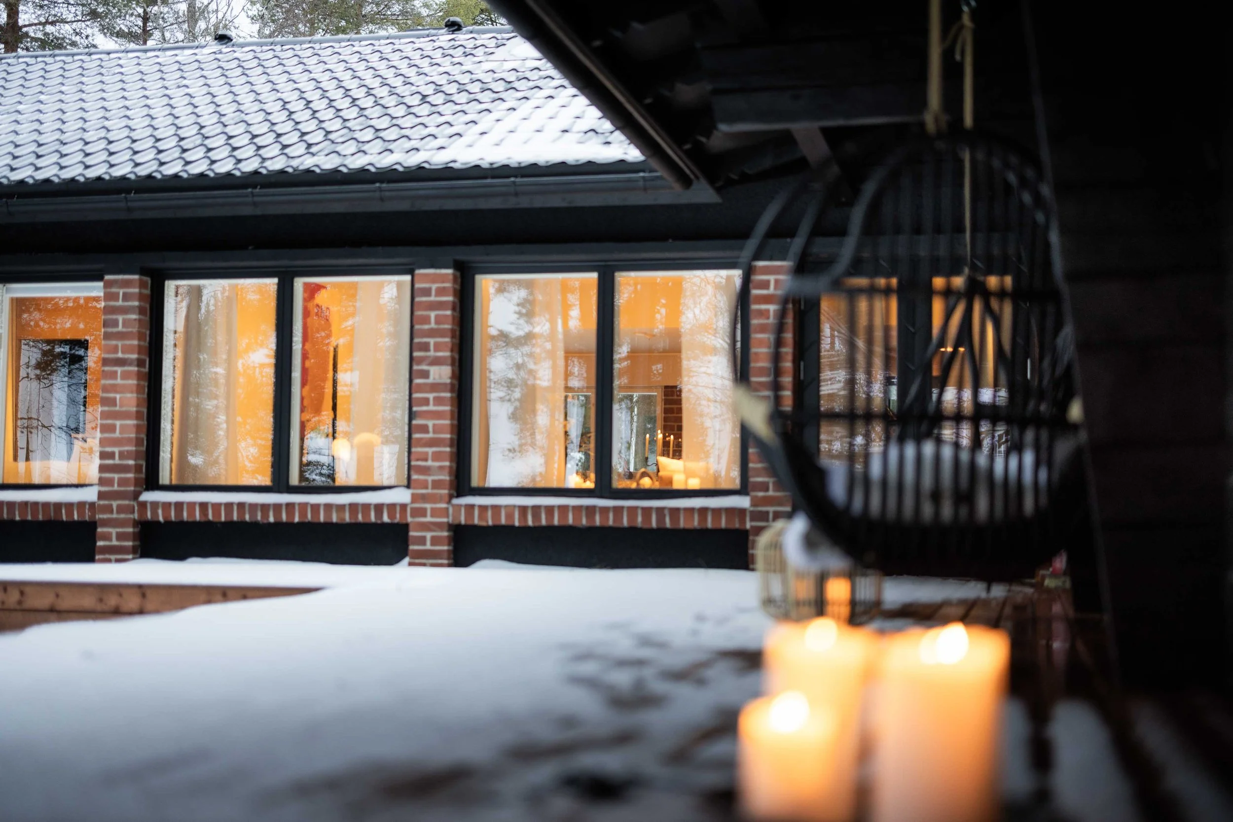 Vista de la casa con ventanas iluminadas en un entorno invernal, vista desde una terraza con velas y una silla colgante
