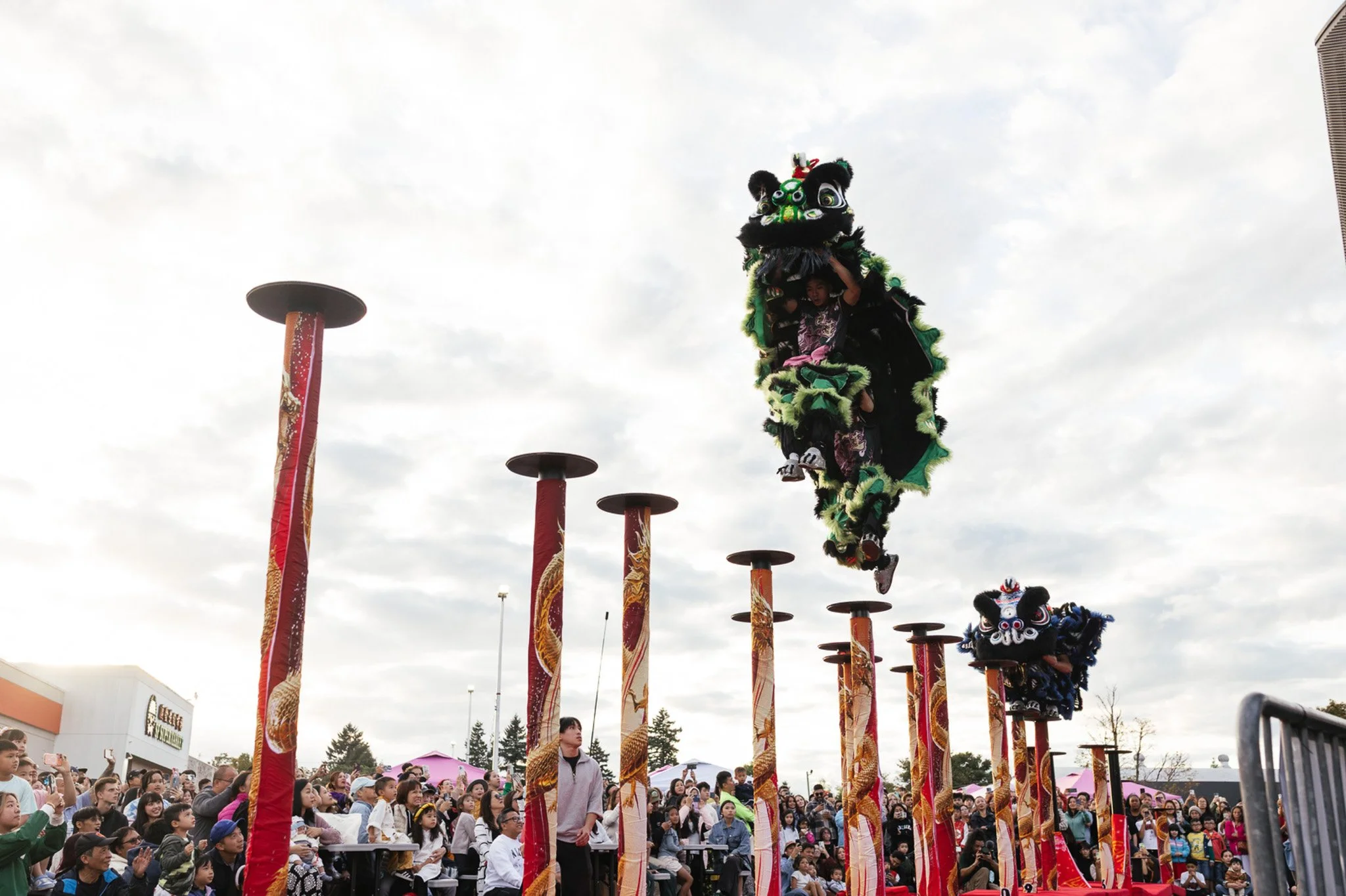 A traditional lion dance performed outdoors during a cultural celebration with a crowd watching. The lion costume is green and black, flying through the air between two performers, with the crowd and cloudy sky in the background.