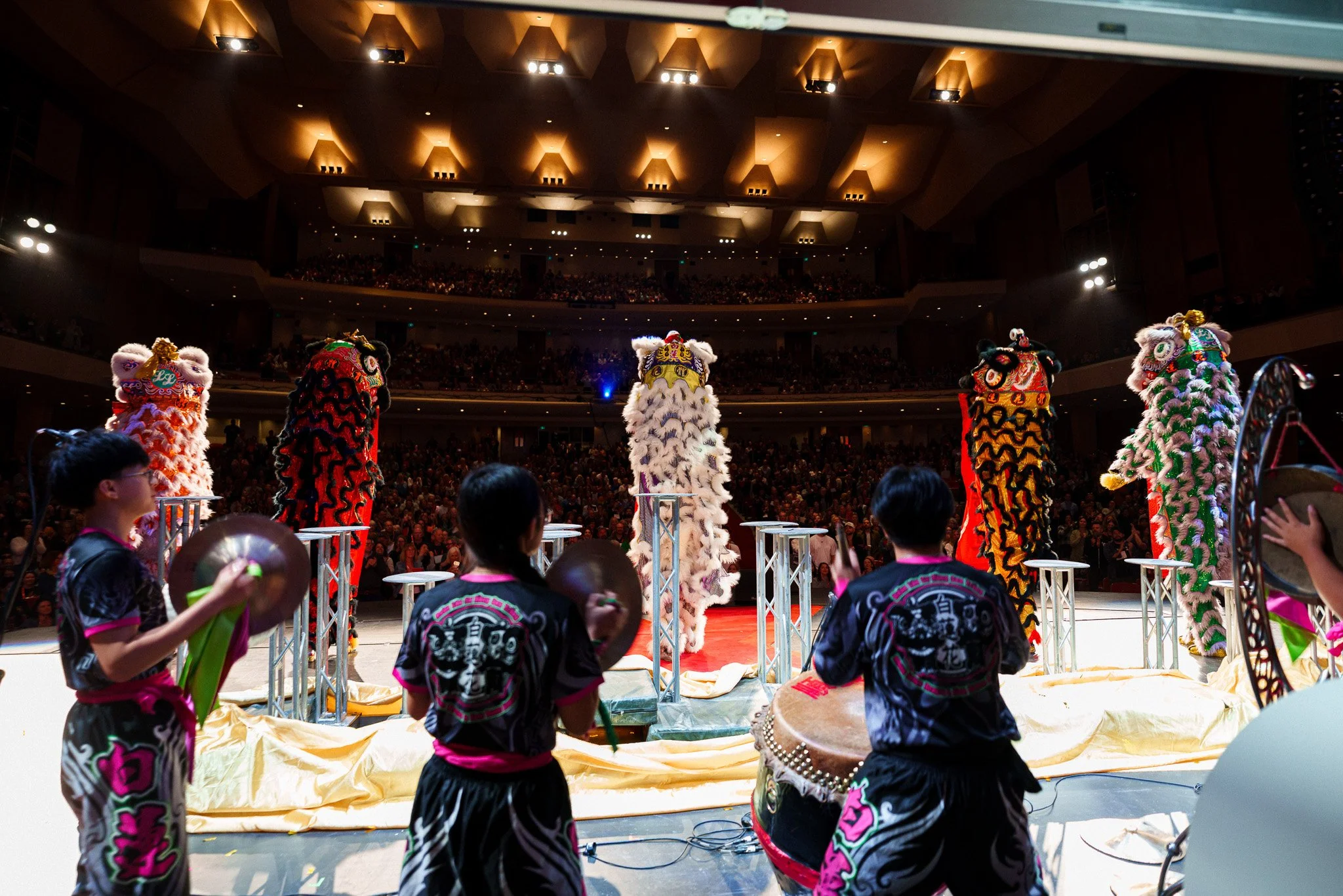 Lion dance performers in colorful costumes on stage with drummers and an audience in the background.