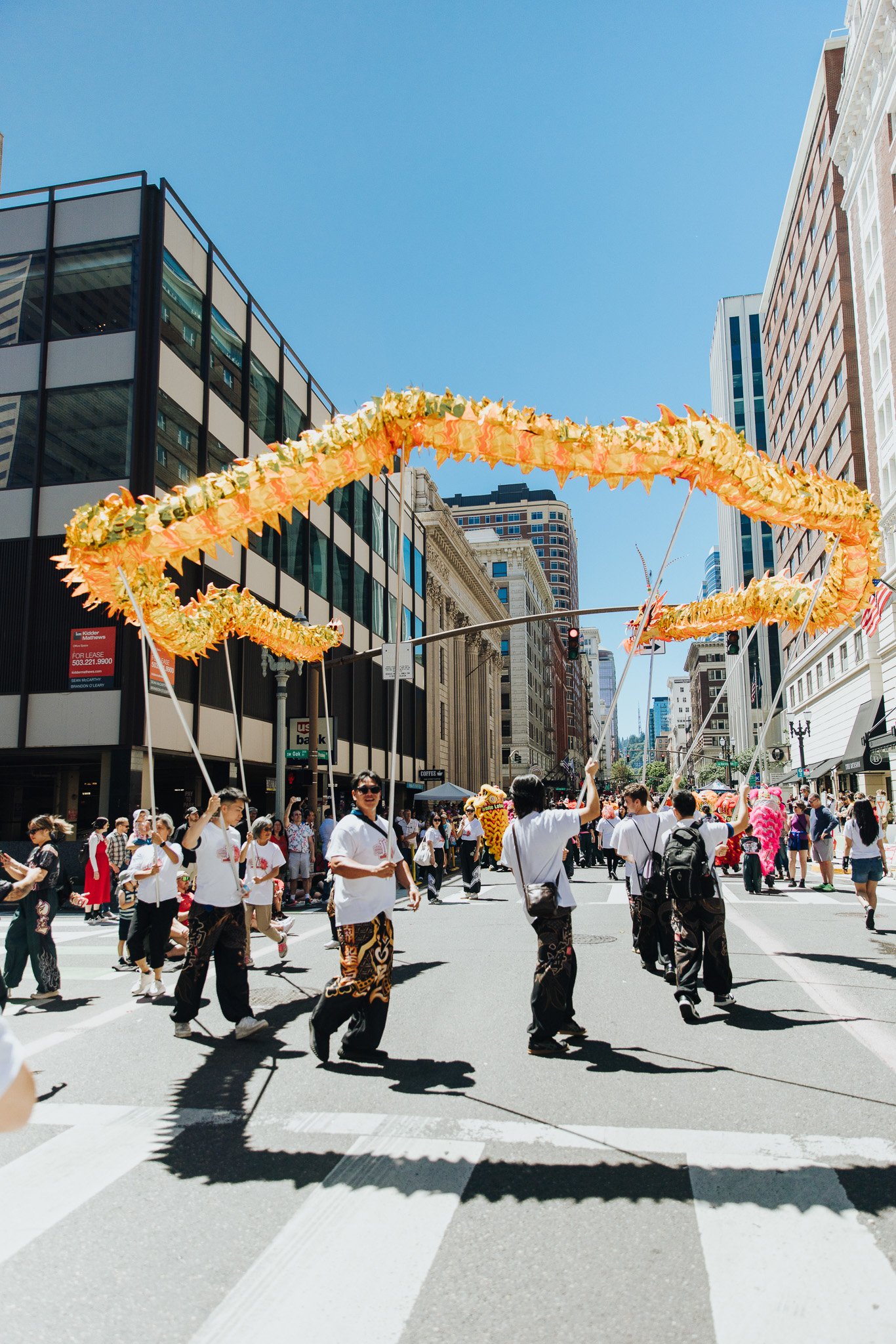 People participating in a parade on a city street, carrying large floral dragon decorations during a celebration, with tall buildings and a clear blue sky in the background.