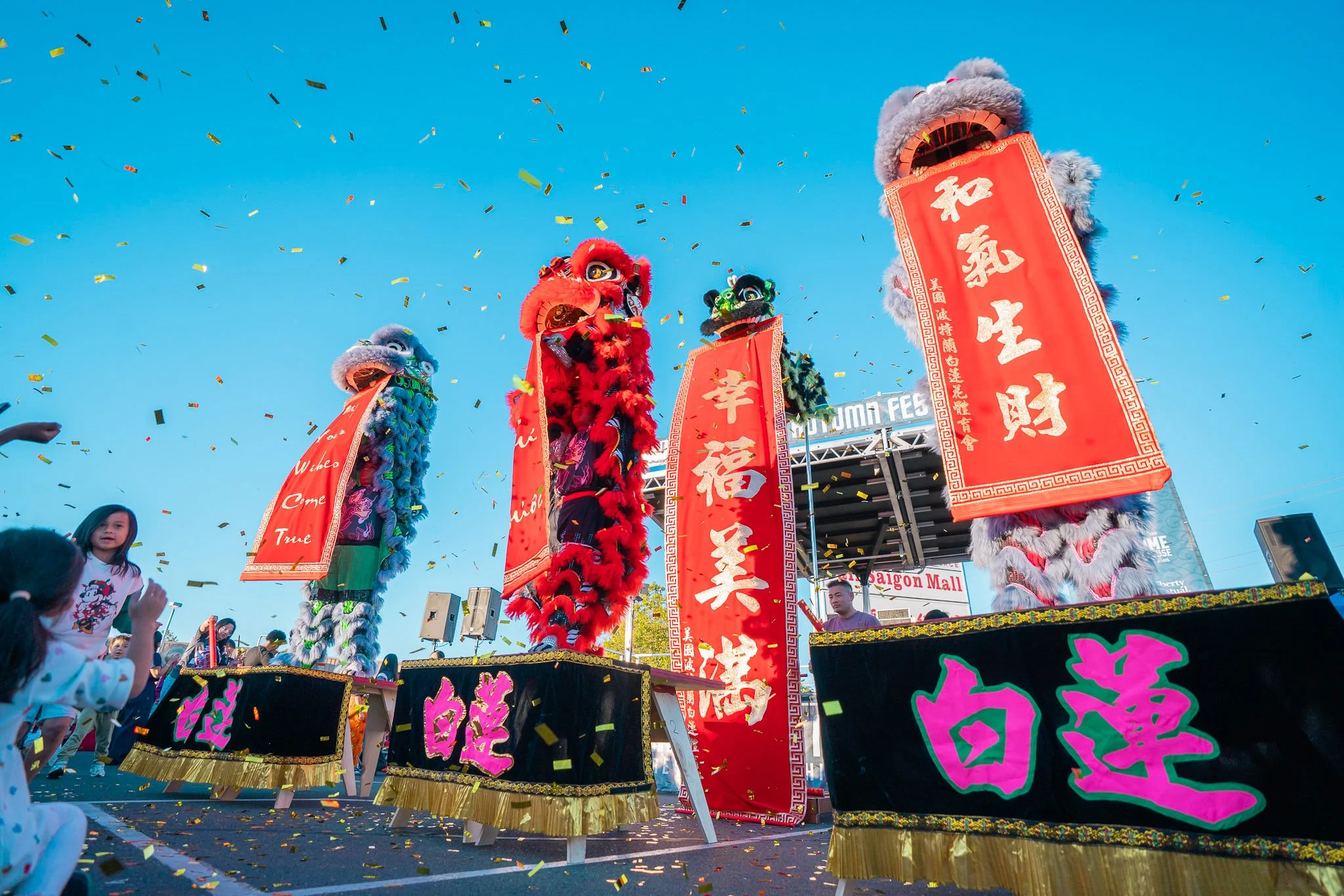 Lion dancers performing during a celebration with colorful costumes, banners, and falling confetti, against a clear blue sky.