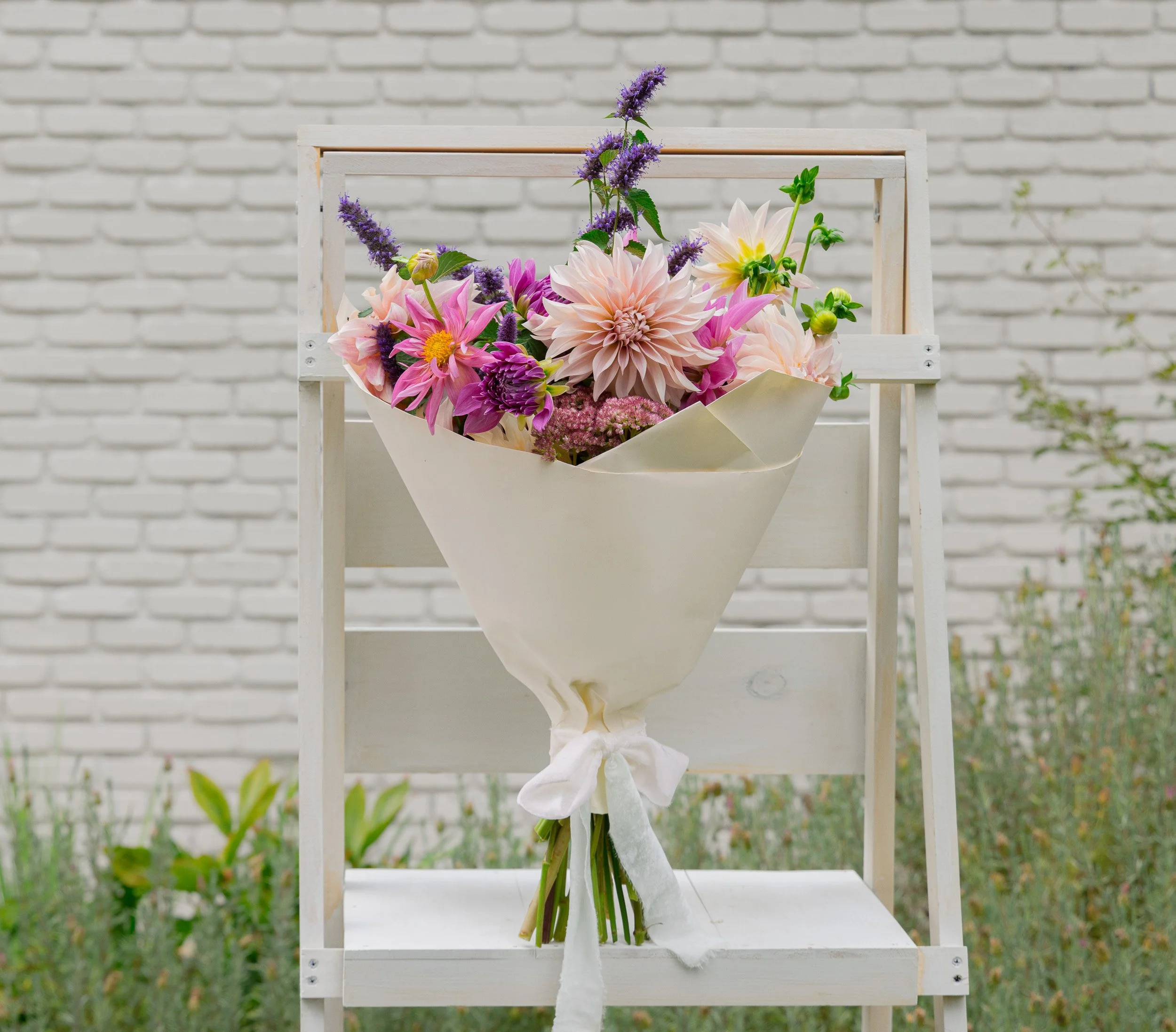 A bouquet of pink and purple flowers with green leaves is displayed in a white wrapping paper, standing on a white wooden stand against a white brick wall.