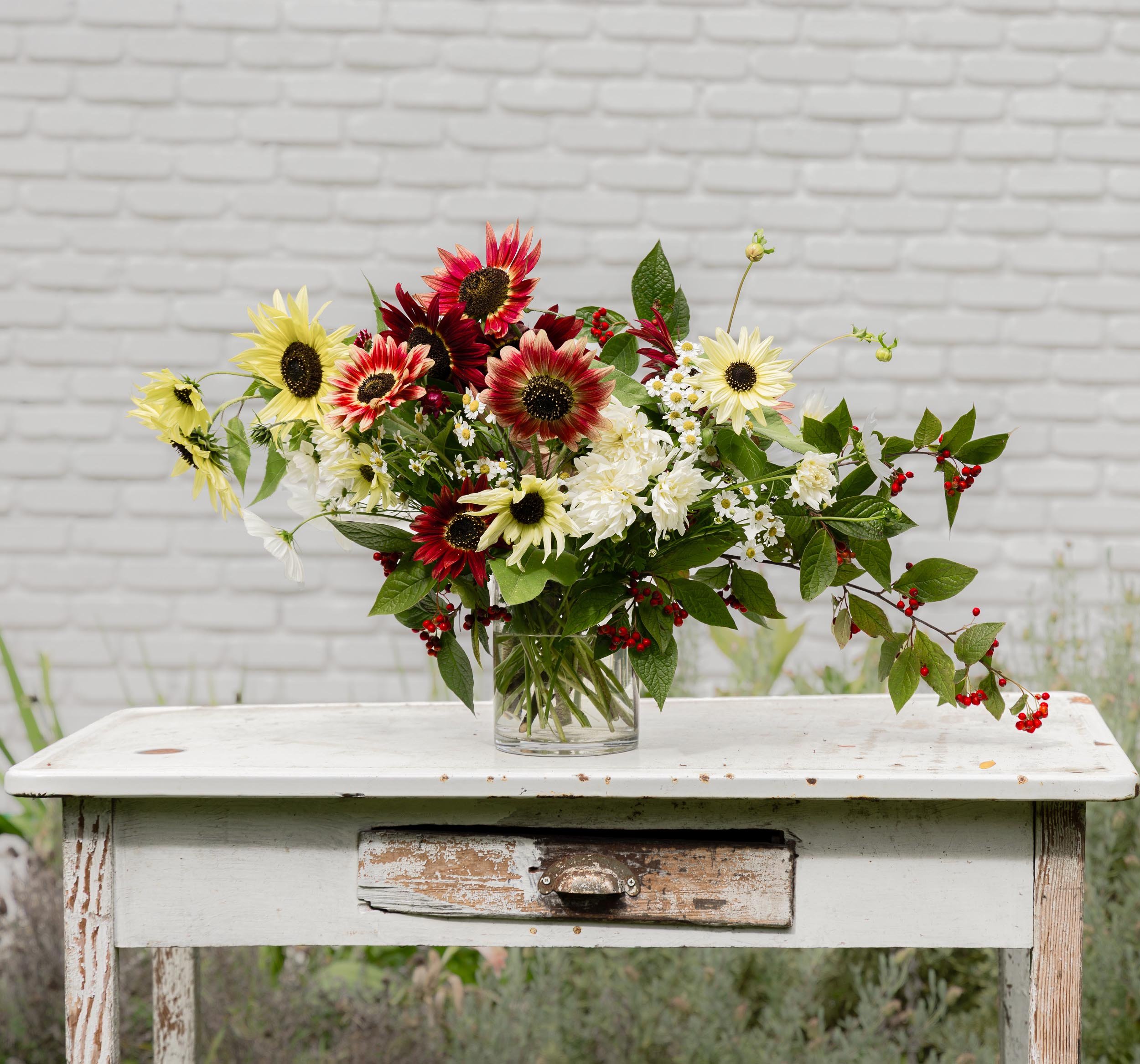 A bouquet of mixed flowers, including yellow and red sunflowers and small white daisies, in a glass vase, placed on a rustic white wooden table with a weathered finish, against a white brick wall background.