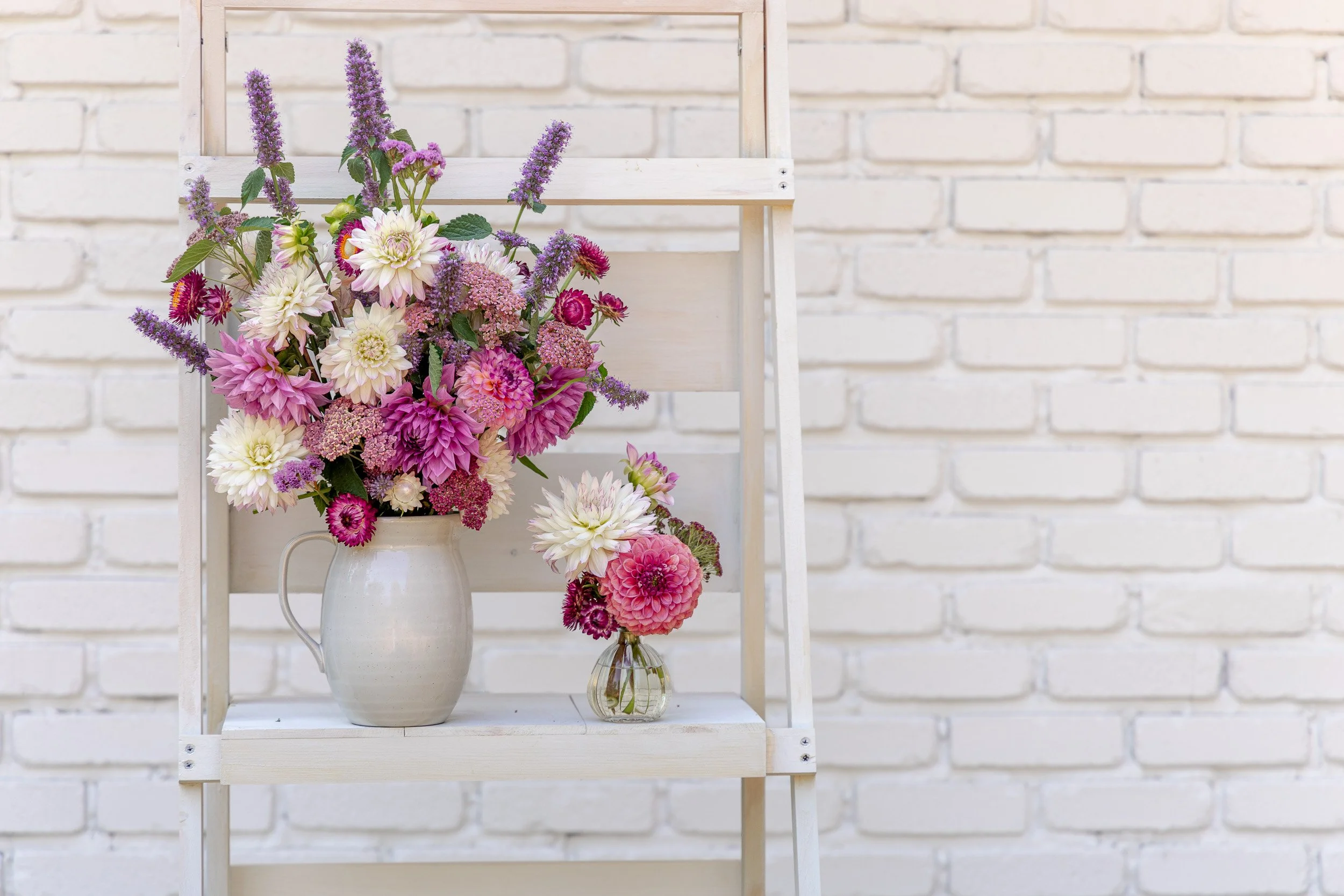A white pitcher and a small glass vase holding a variety of pink, purple, and white flowers, placed on a white wooden shelf against a white brick wall.