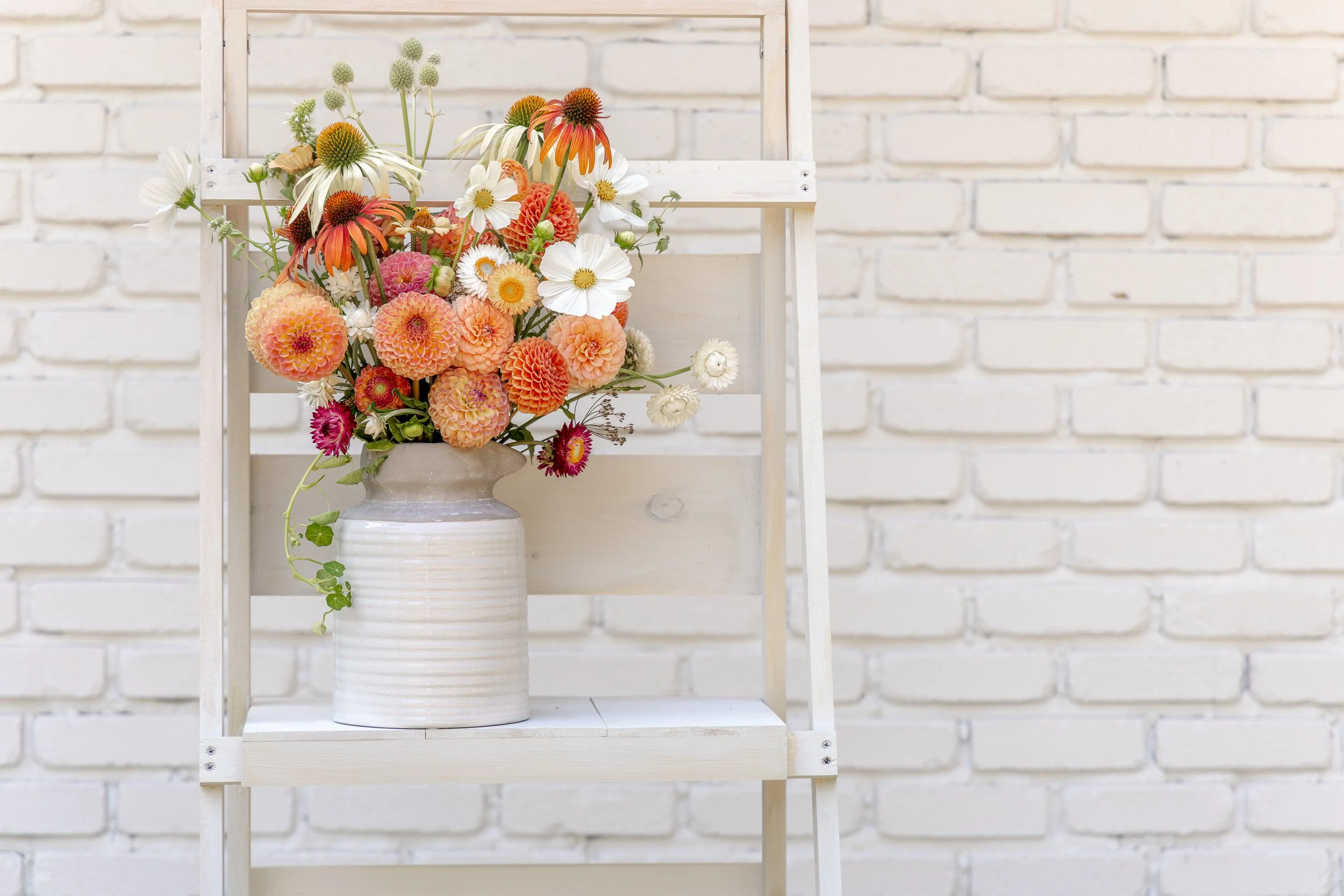 A white vase filled with a colorful arrangement of flowers including orange, white, pink, and cream-colored blossoms, placed on a white shelf against a white brick wall.