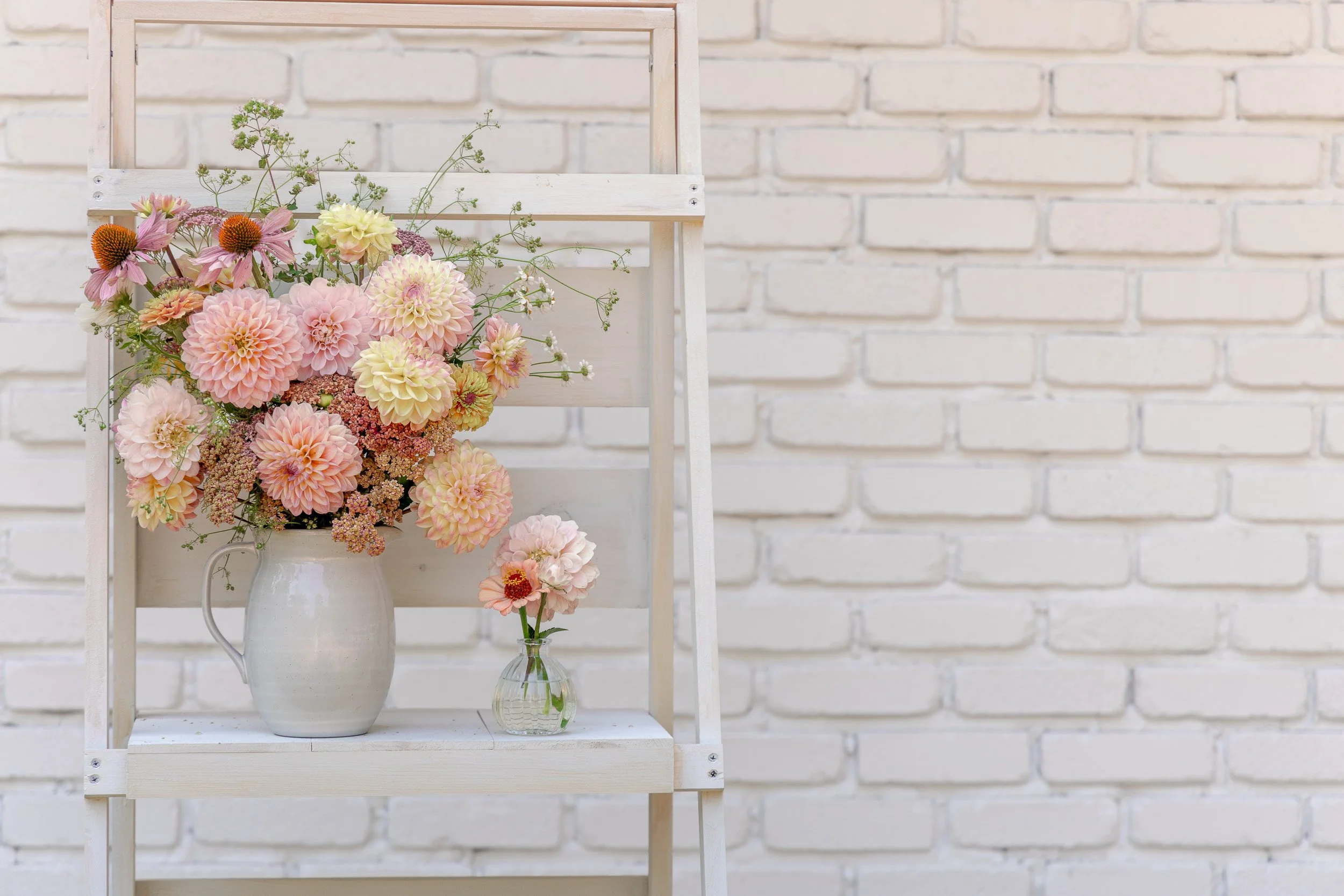 A white pitcher and a small clear glass vase with flowers sit on a white shelf against a white brick wall. The pitcher is filled with a bouquet of pink, peach, and cream-colored flowers with various greenery.